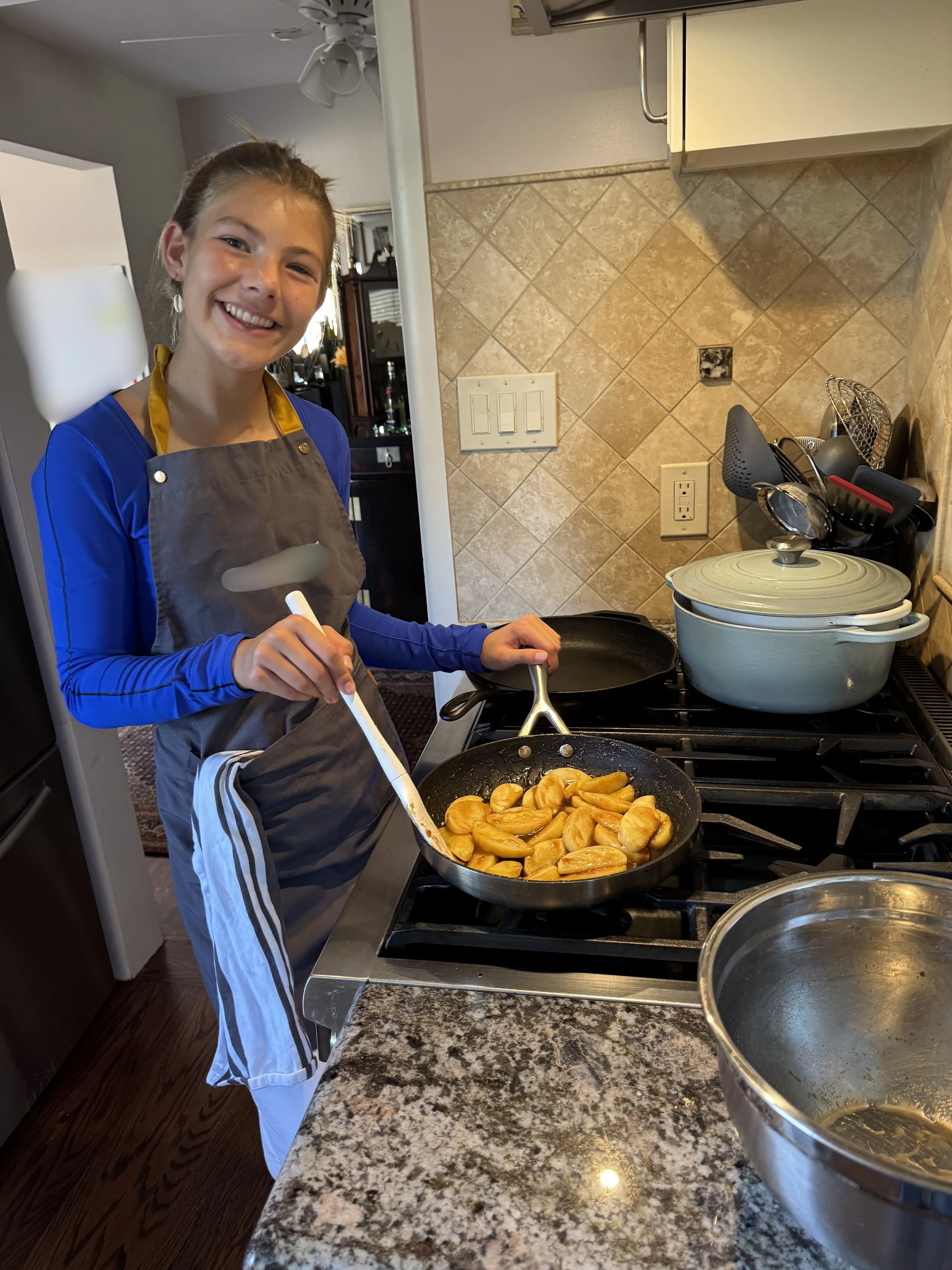A girl smiling while cooking on a stovetop in a kitchen, preparing sliced bananas in a frying pan.