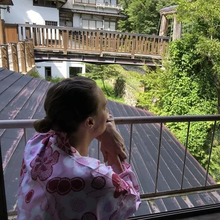 A woman standing on a balcony, gazing out at a scenic view with trees and wooden buildings, wearing a pink and white floral-patterned garment.