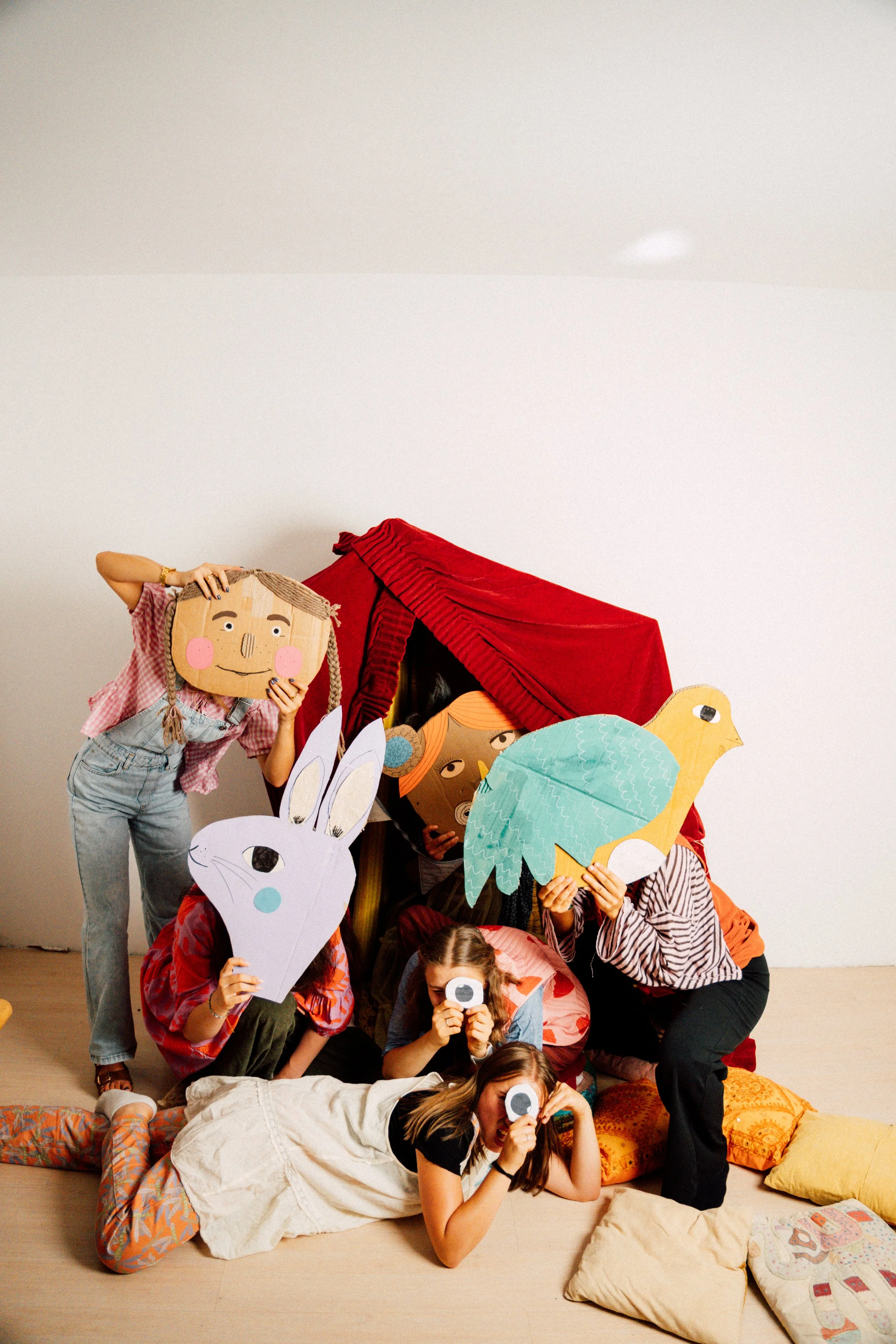 Gruppe von Kindern bei einer Puppentheater-Aufführung mit Tier- und Figurenmasken, die auf dem Boden sitzen, während andere Kinder und Erwachsene die Bühne im Hintergrund halten.