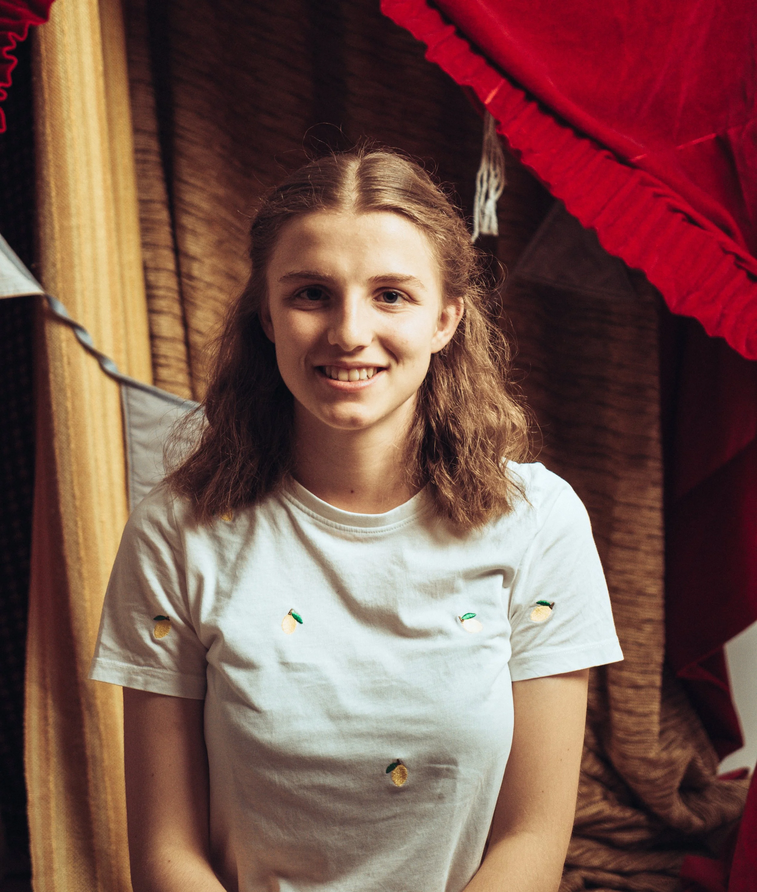 Nahaufnahme eines lachenden jungen Mädchens mit braunen, gewellten Haaren, das weiße T-Shirt mit kleinen Eiscreme-Emojis trägt, im Hintergrund bunte Stoffe.