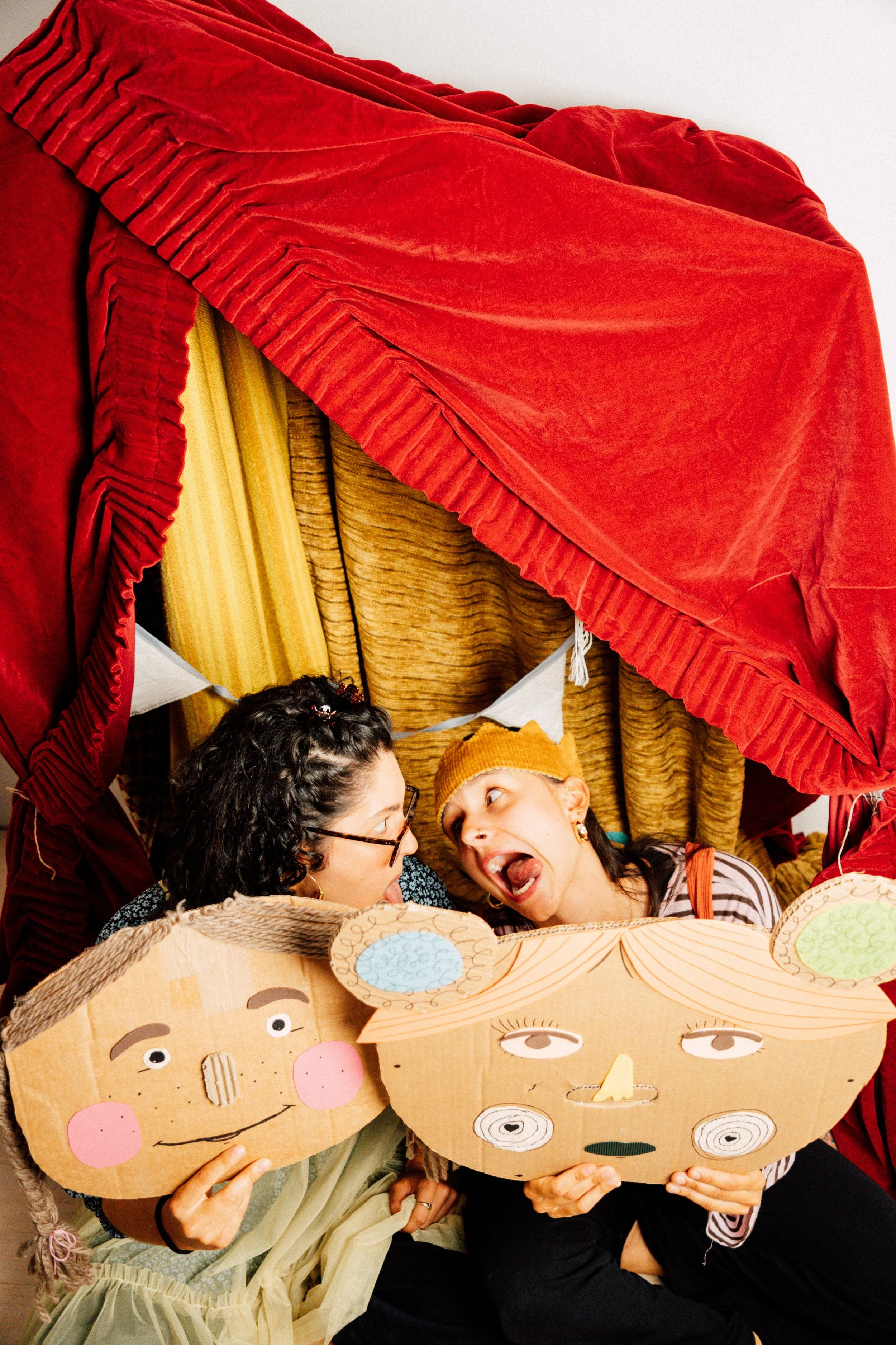 Zwei Frauen halten große Papiermasken mit lachenden Gesichtern, während sie in einem Raum sitzen. Im Hintergrund sind bunte Vorhänge und eine Person, die mit einer roten Decke oder Gardine spielt.