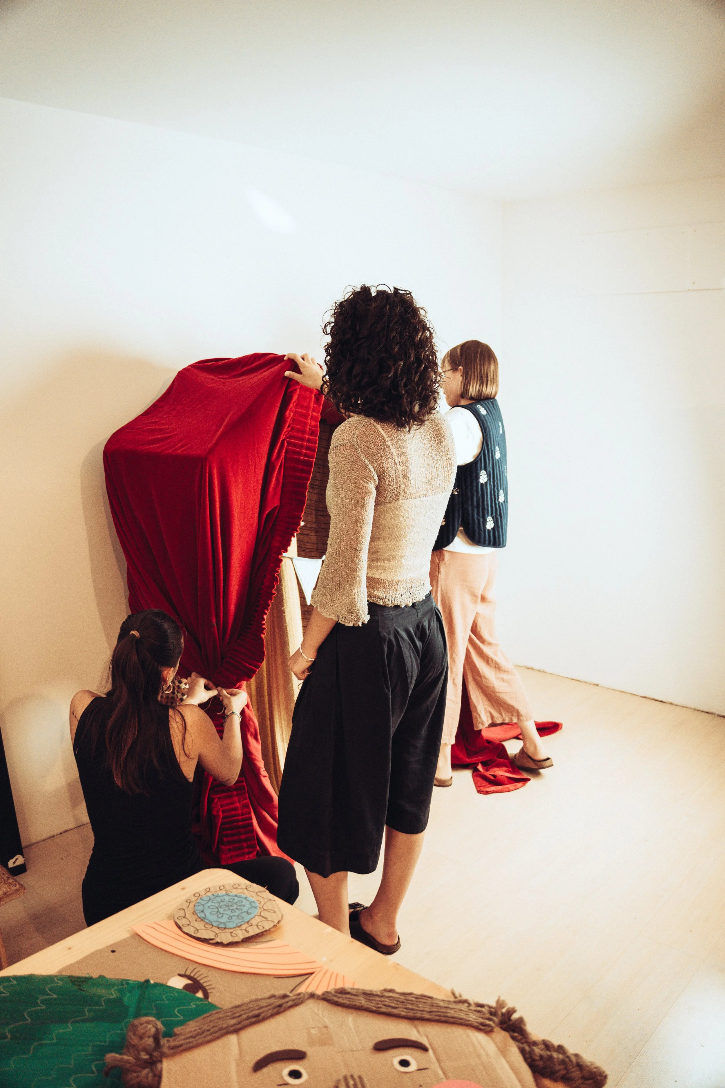 Mehrere Menschen in einem Raum, einige steigen hinter einem roten Vorhang hervor, der auf einem Ständer hängt, andere schauen zu. Es gibt verschiedene Kunstwerke und dekorative Gegenstände im Vordergrund.
