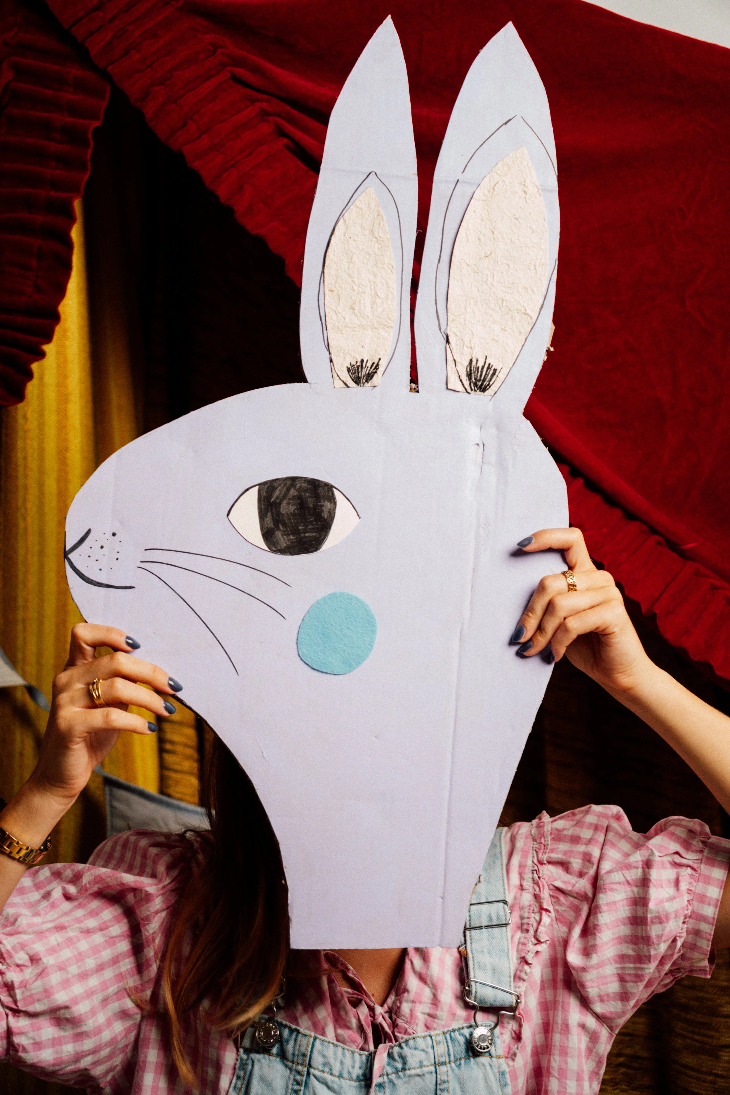 Person hält ein handgemachtes Osterhasenkopf-Pappmasken mit langen Ohren und einem großen schwarzen Auge, auf rotem und gelbem Stoffvorhängen im Hintergrund.