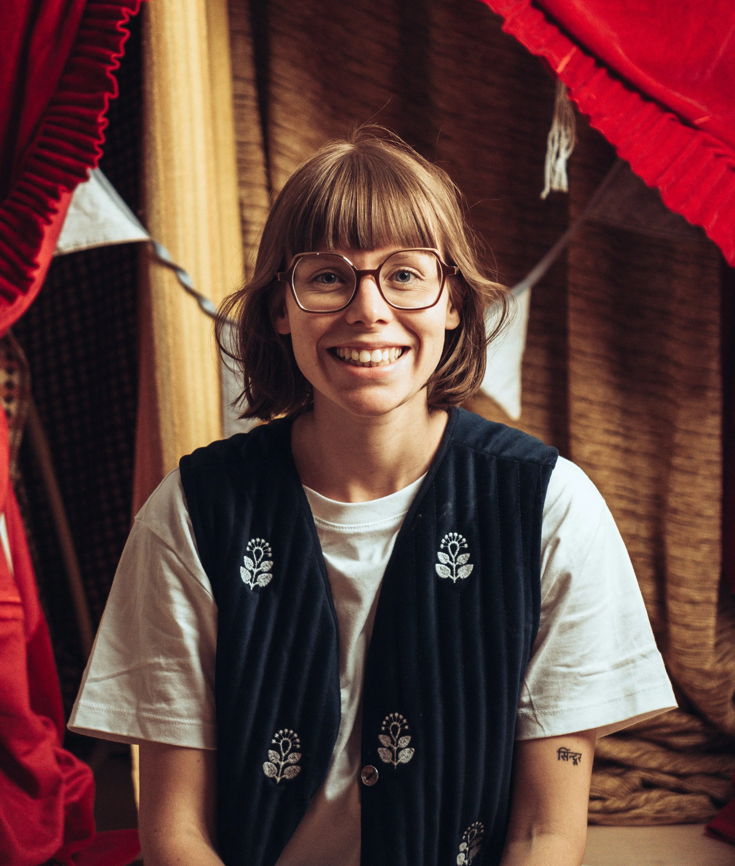 Junge Frau mit Brille, lachend, in traditioneller Kleidung, neben roten und gelben Stoffen, Tätowierung auf dem Arm.