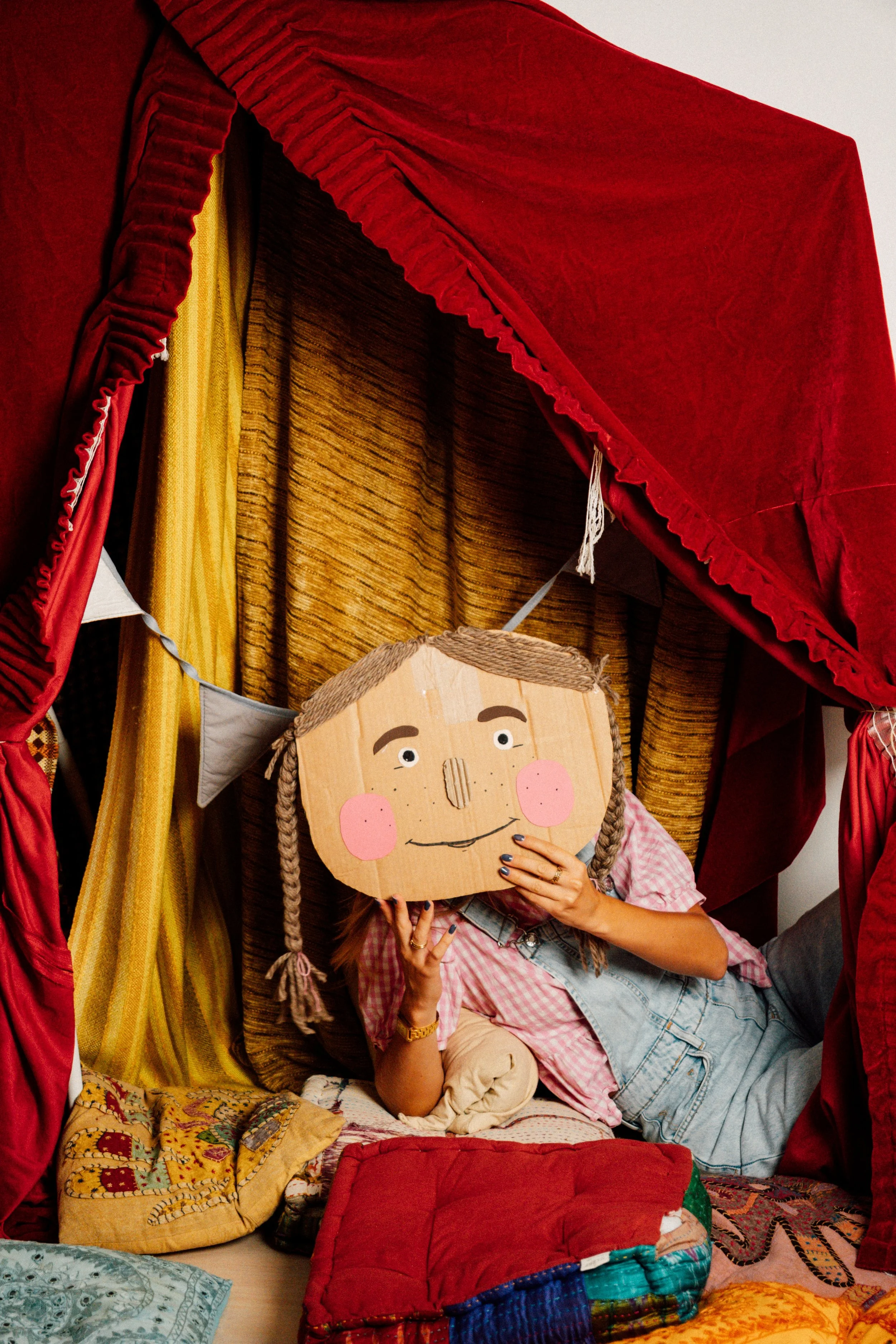 Person hält eine handgemachte Pappfigur mit einem lachenden Gesicht vor das Gesicht, im Hintergrund bunte Stoffe und Kissen. Die Person trägt ein gestreiftes Hemd.