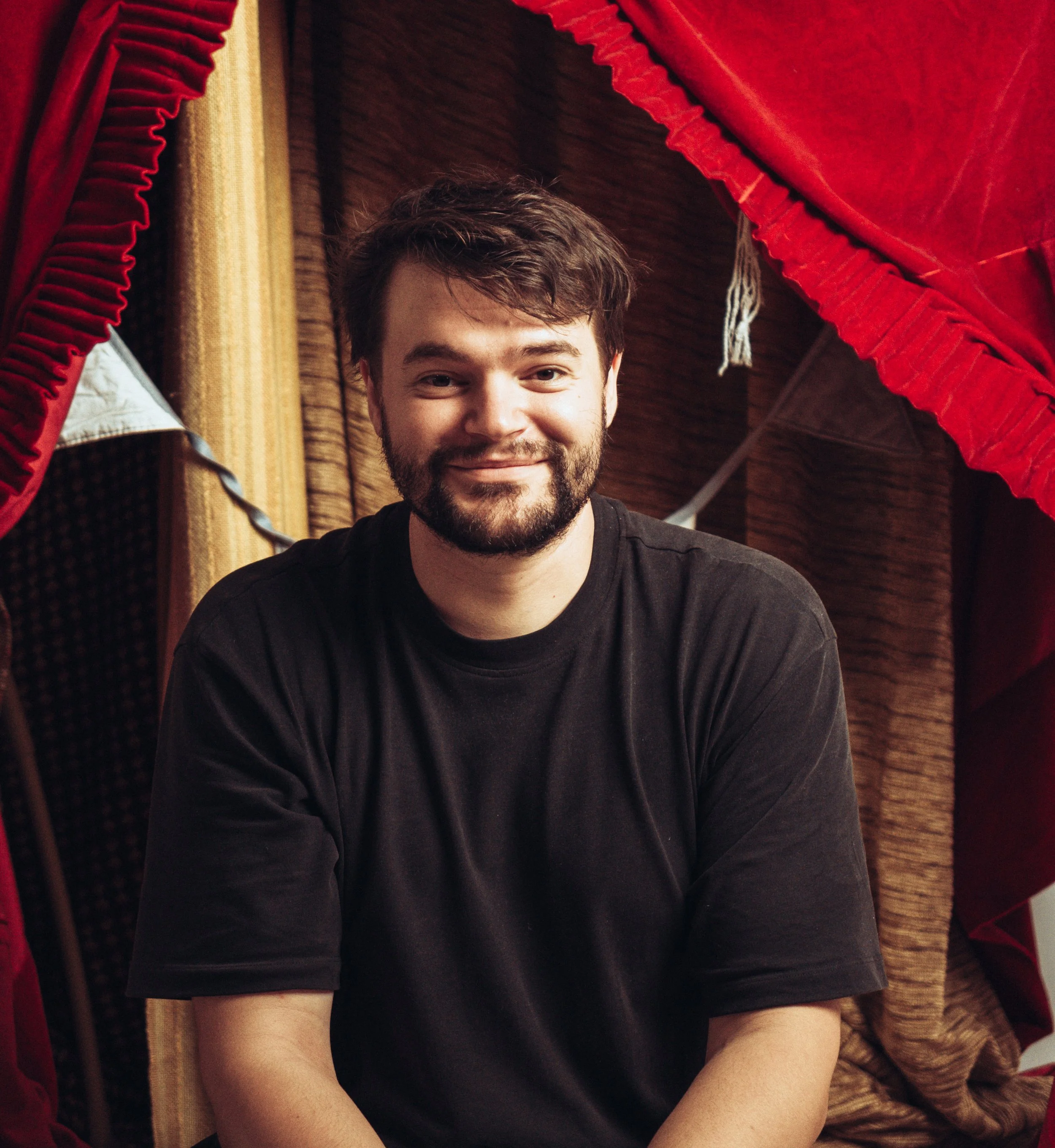 Ein Mann mit braunem, gewelltem Haar und Bart trägt ein schwarzes T-Shirt. Er sitzt vor einem Hintergrund aus verschiedenen bunten Vorhängen, darunter rot, gelb und braun, die an einem Holzbalken hängen.