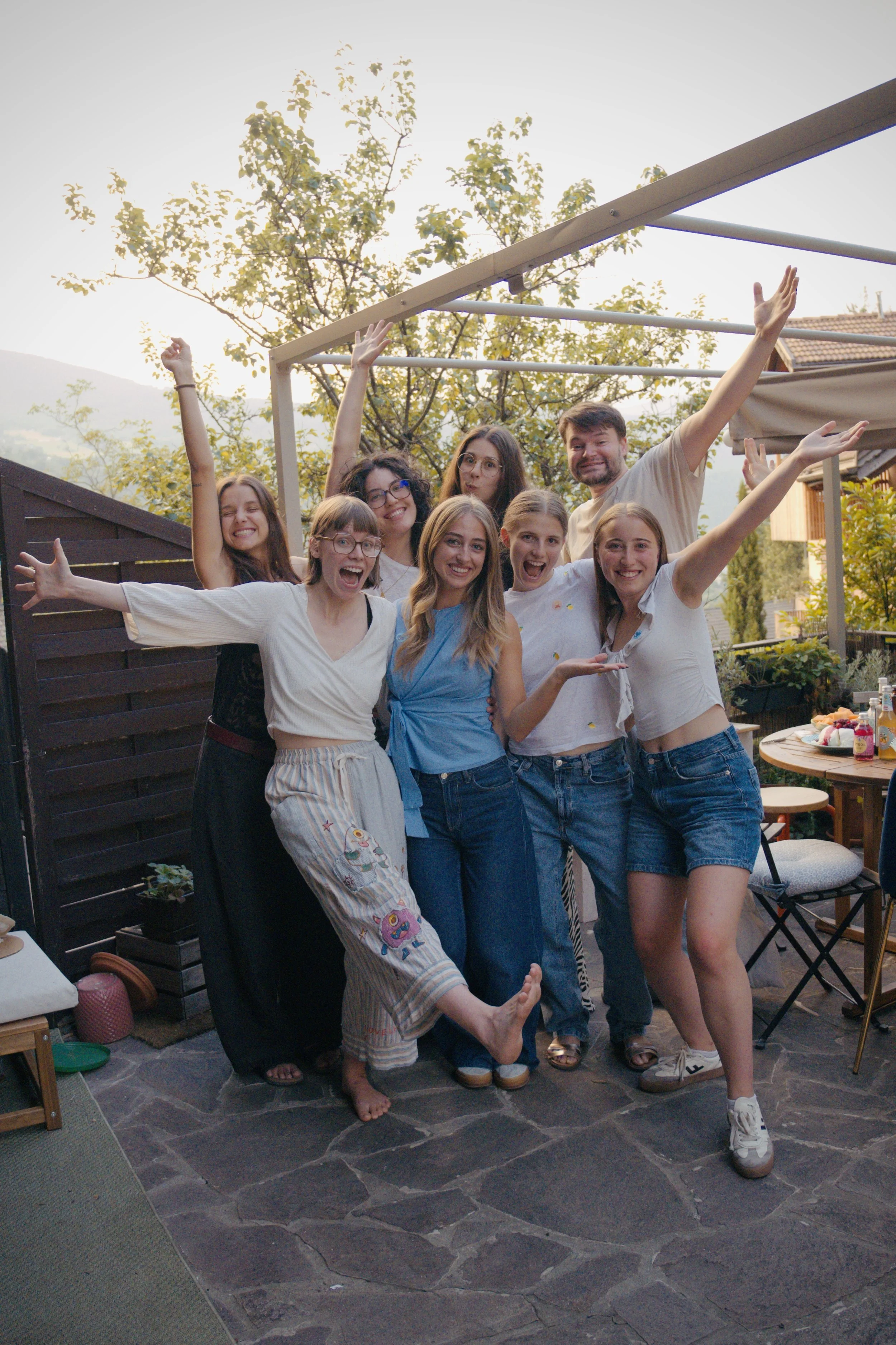 Eine Gruppe junger Menschen, die auf einer Terrasse im Freien fröhlich zusammen feiern und lachen, umgeben von Bäumen und einem blauen Himmel.