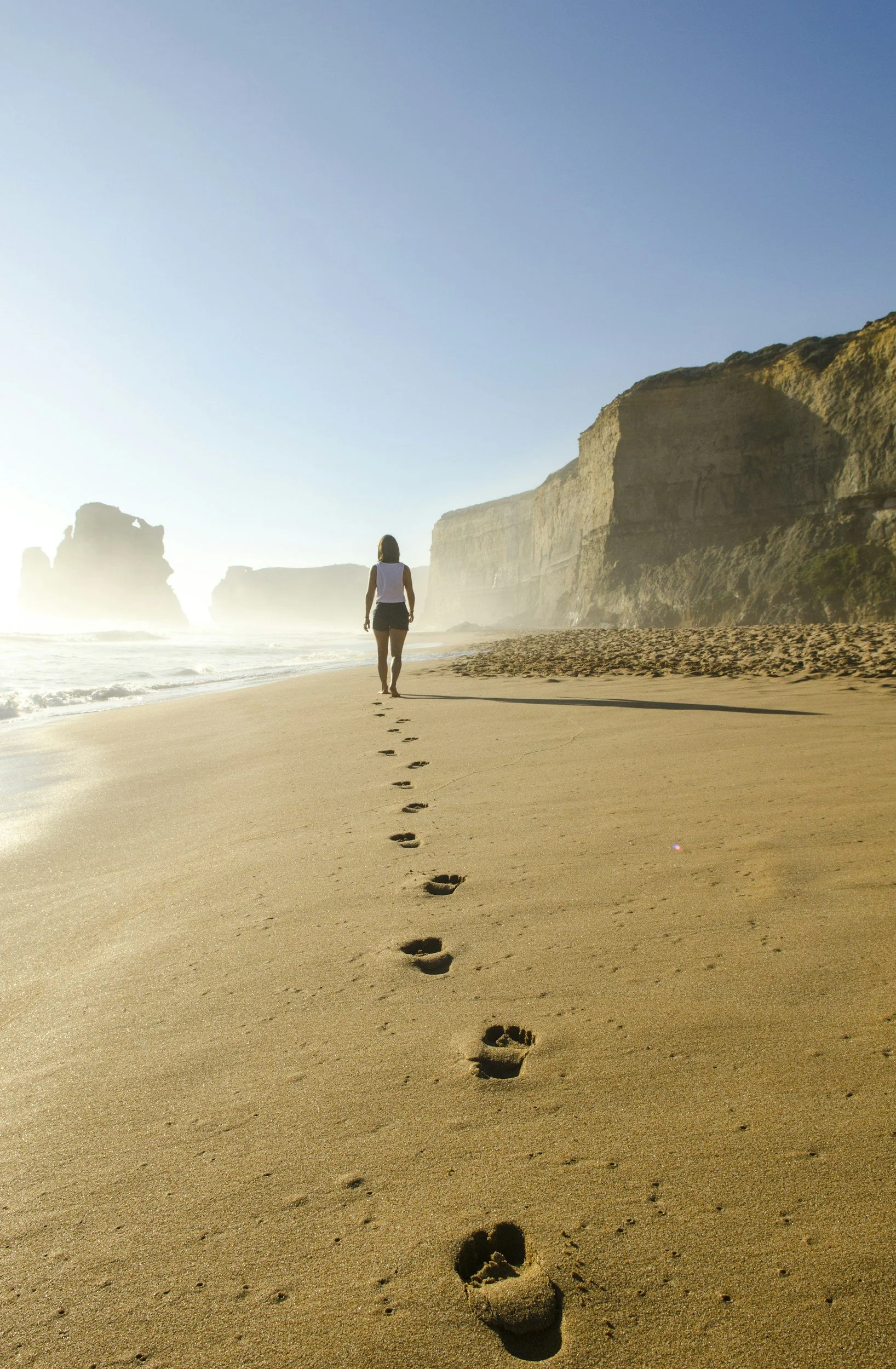 A person walking along a sandy beach with footprints trailing behind, towering cliffs on the right, and the ocean on the left under a clear blue sky.