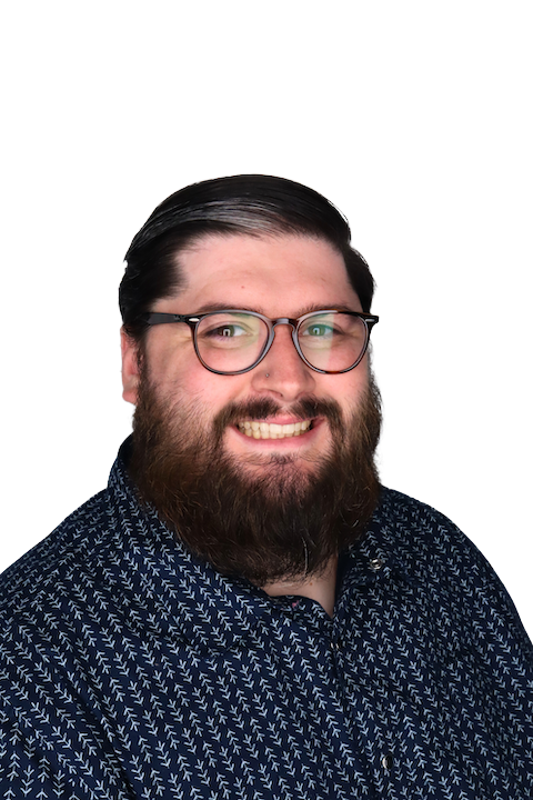 A man with glasses and a beard smiling, wearing a dark patterned shirt, against a plain black background.