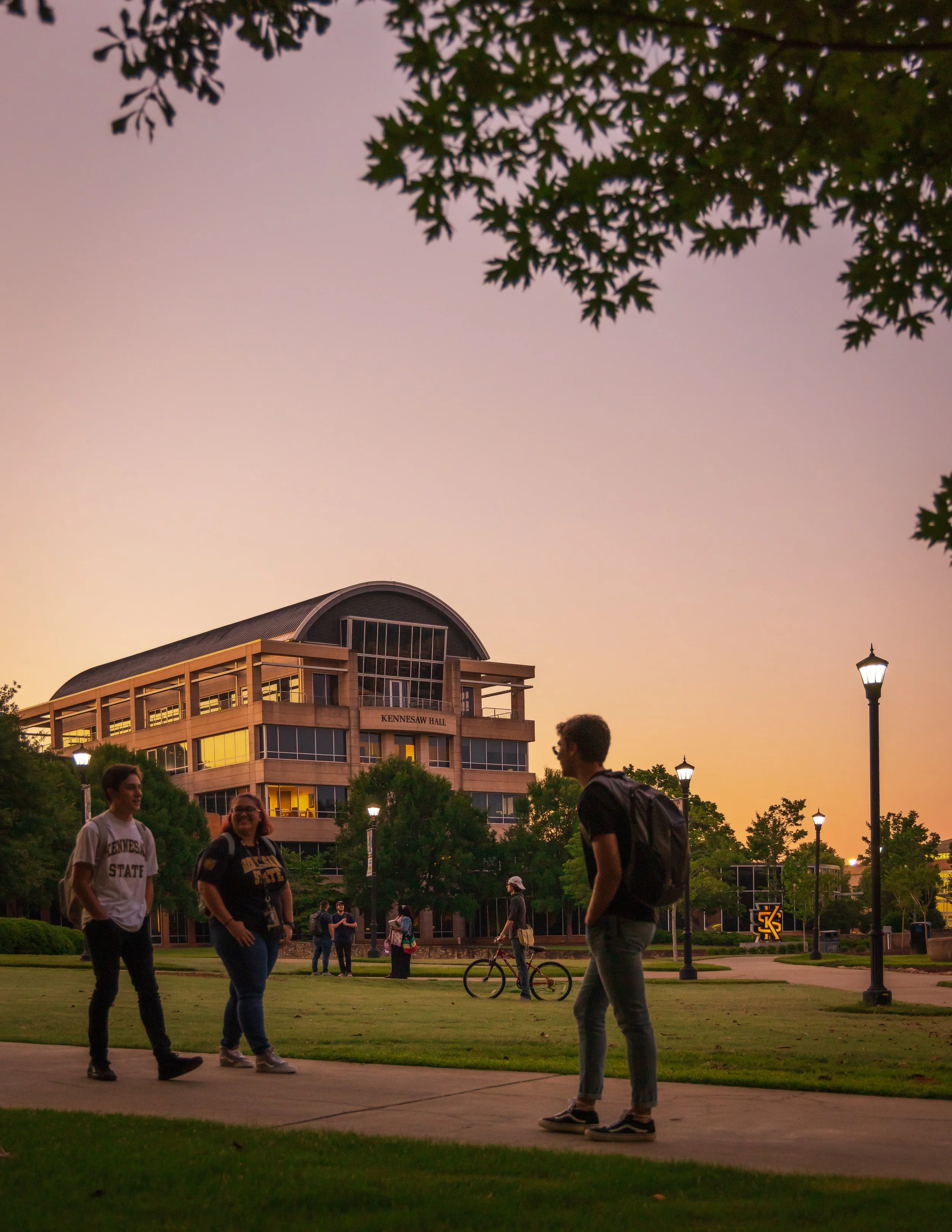 Group of students walking and talking on a campus lawn during sunset. In the background, a building labeled 'Kennesaw Hall' and street lamps are visible.
