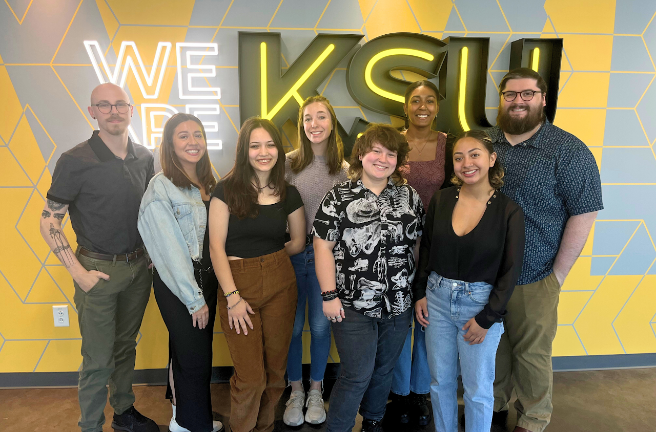 Group of 8 student and faculty researchers standing in front of a lighted sign that says We Are KSU