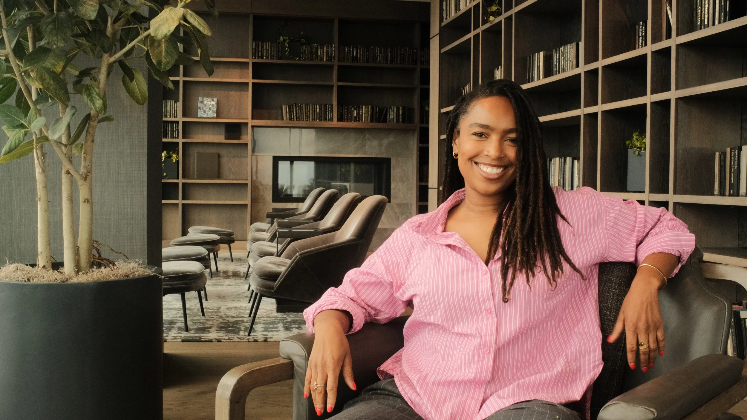 A woman with long dreadlocks wearing a pink striped shirt smiling while sitting in a modern library or lounge area with black chairs, bookshelves, and a large potted plant.
