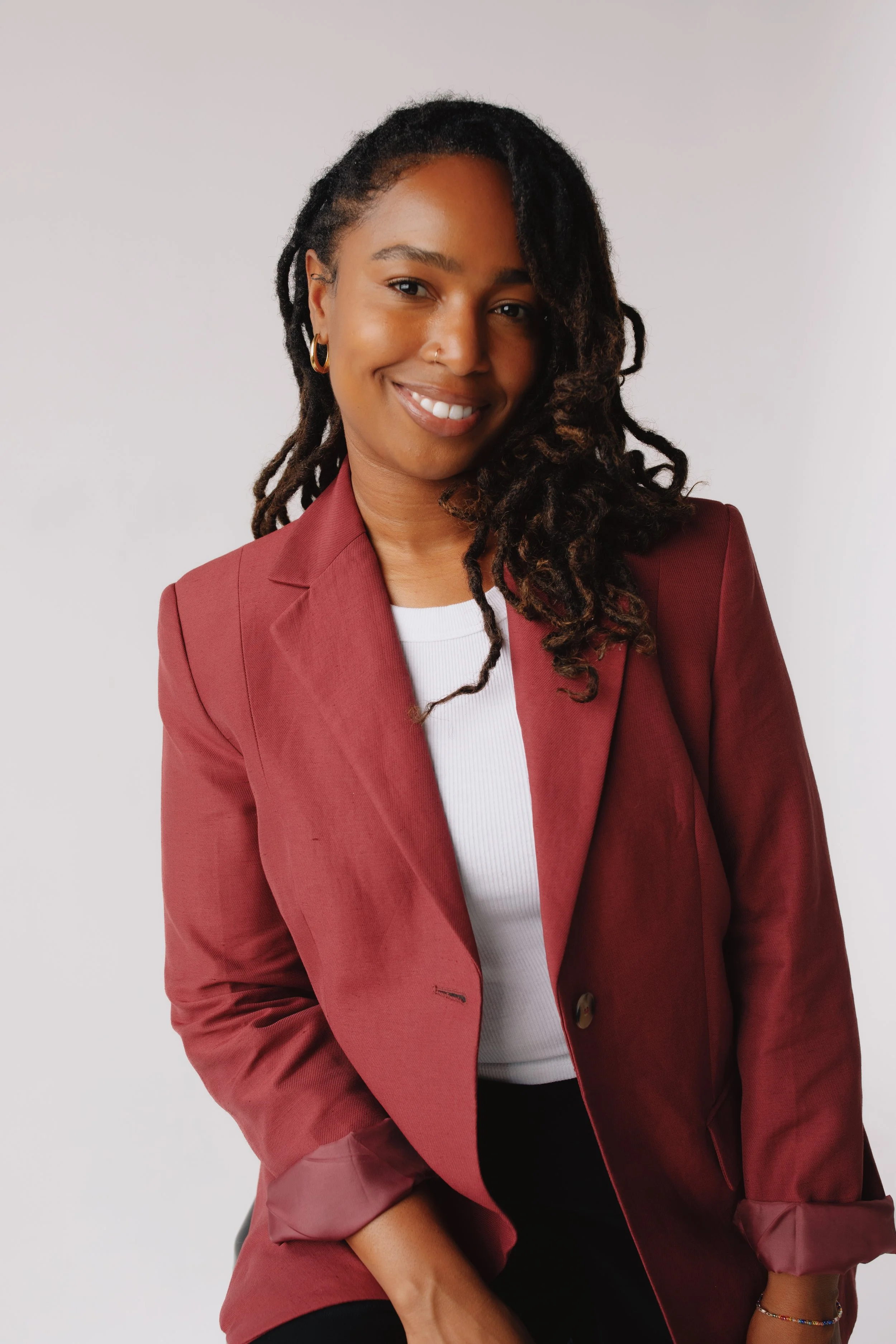 A woman with dark curly hair wearing gold hoop earrings, a nose ring, a maroon blazer, and a white shirt, smiling against a plain light background.
