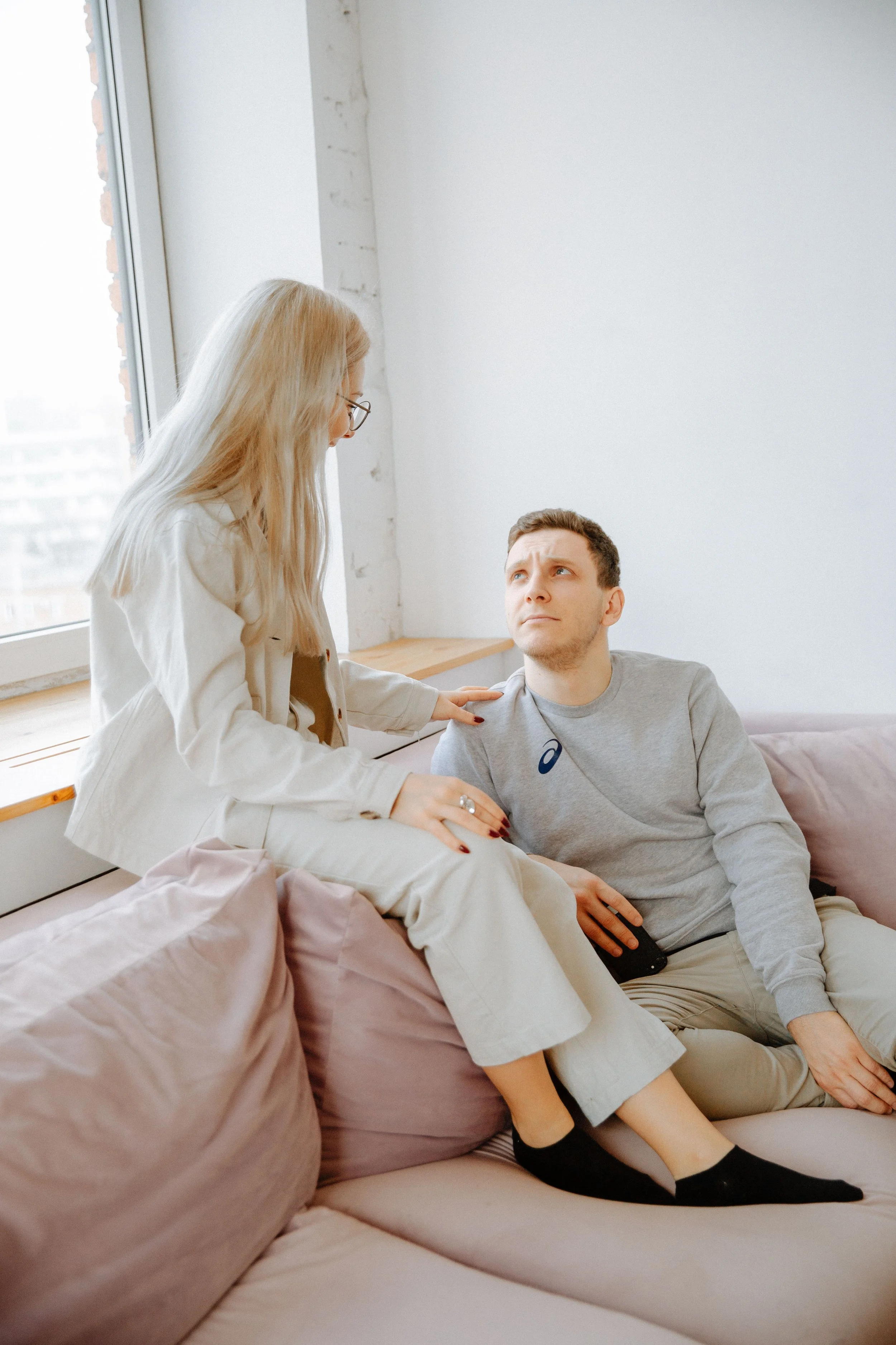 A woman with blonde hair and glasses is sitting on a beige couch, leaning towards a man with short hair who is seated, holding a smartphone in his hand. They are in a bright room with a large window and white walls.