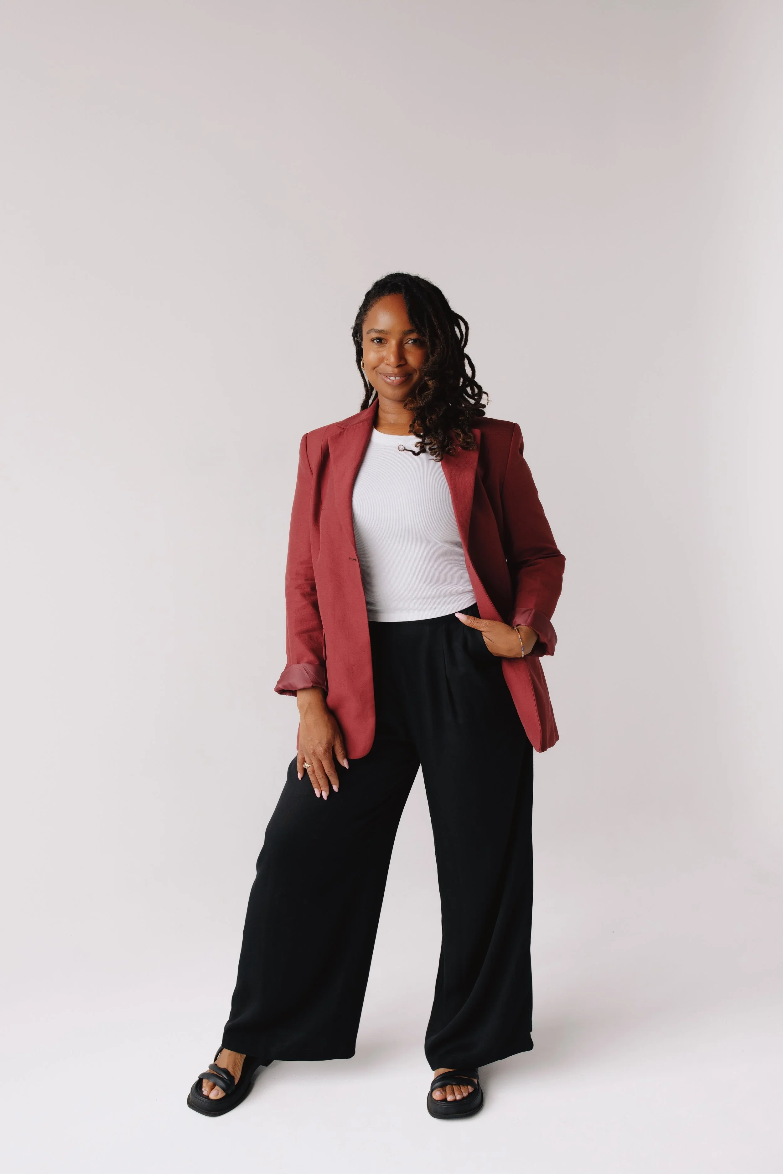 A woman with dark curly hair and medium skin tone posing against a plain white background, wearing a white t-shirt, maroon blazer, black wide-leg pants, and black sandals.