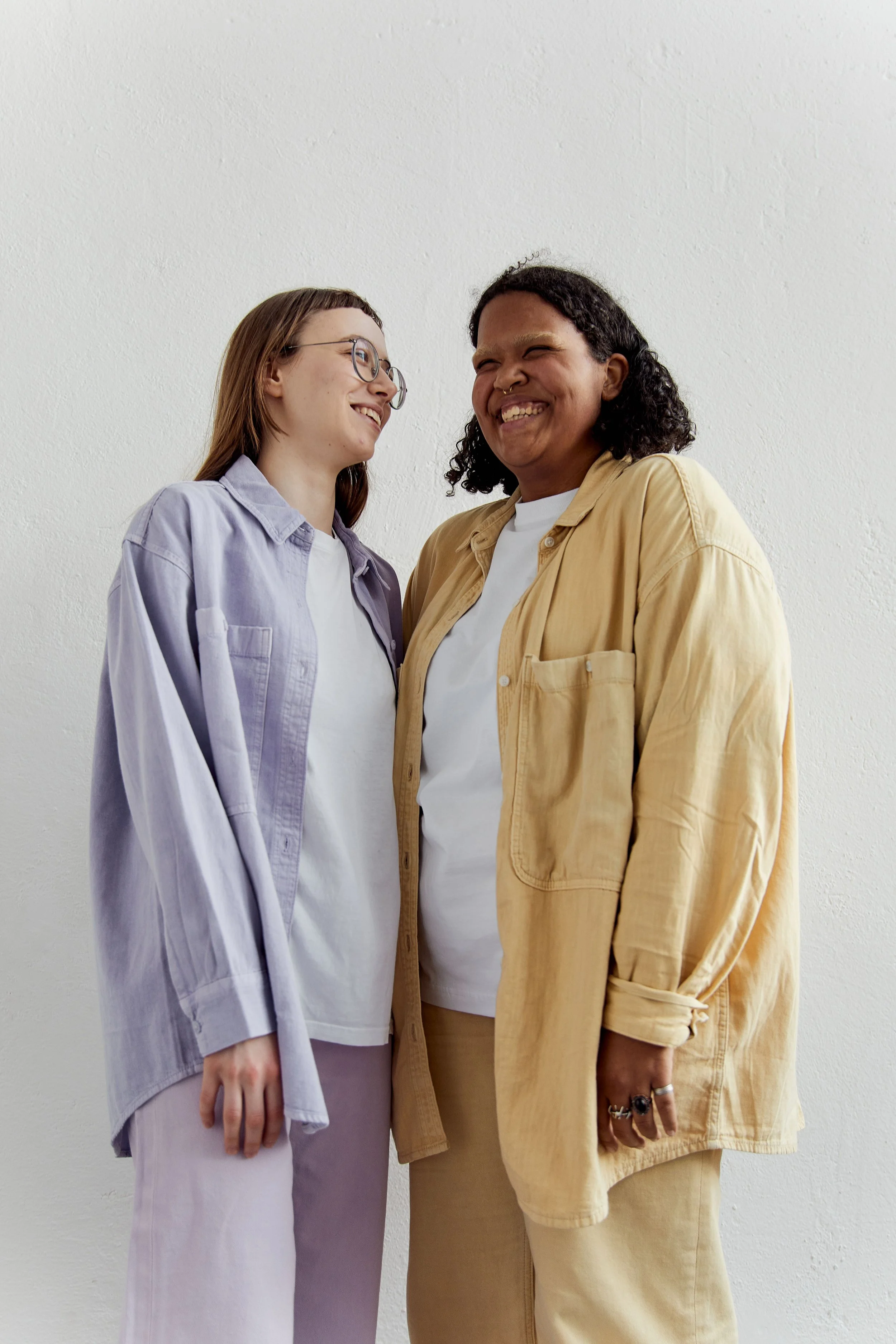Two women standing close together against a white wall, smiling and looking at each other. One woman has glasses and light brown hair, wearing a light purple jacket and pants. The other has dark curly hair, wearing a beige shirt and matching pants, with rings on her fingers.