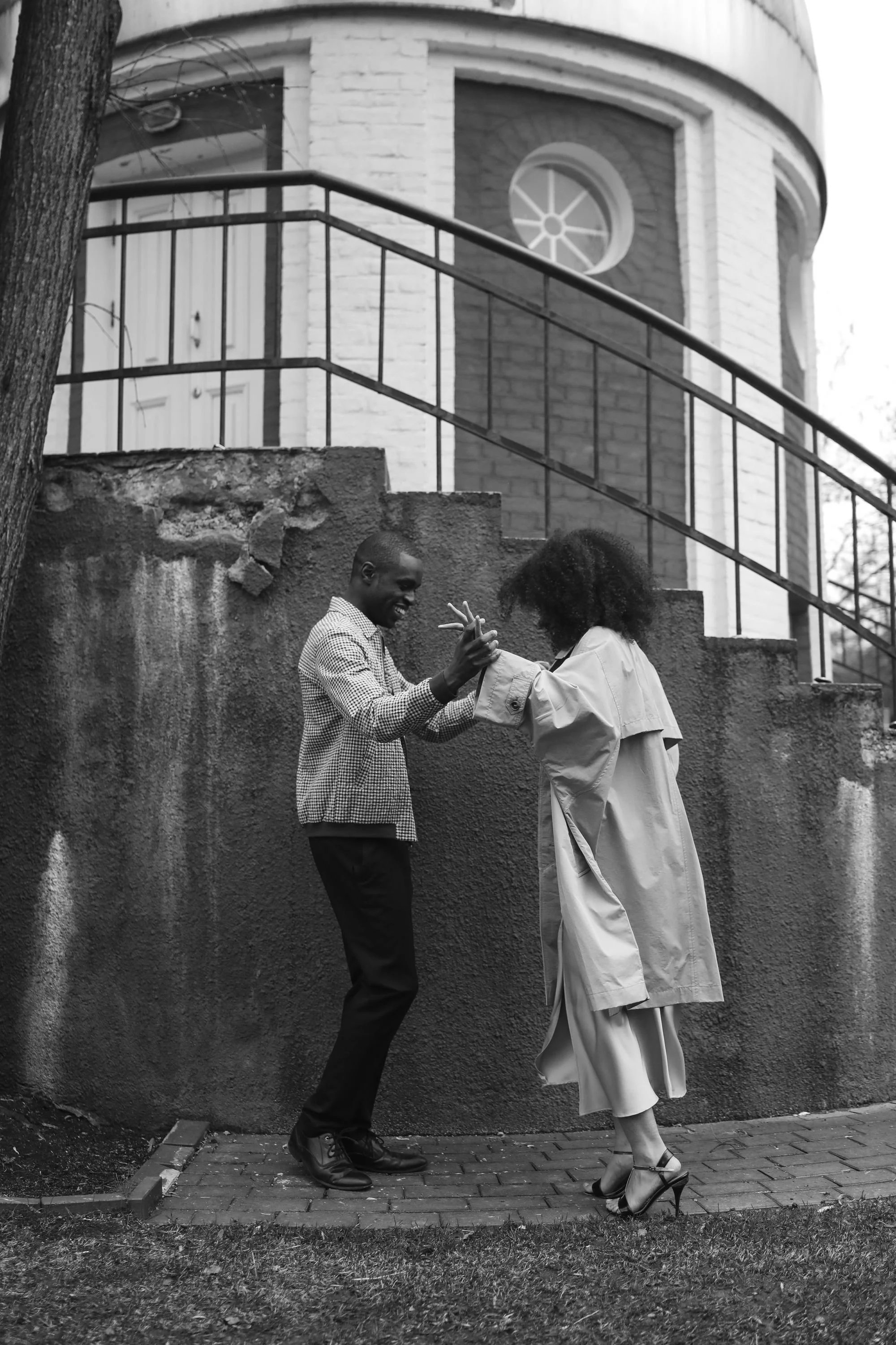 A man and woman dancing together outdoors on a sidewalk in front of a brick wall and house with a railing and circular window. The man is smiling and holding the woman's hand, while the woman, wearing high heels and a trench coat, is leaning back gracefully.