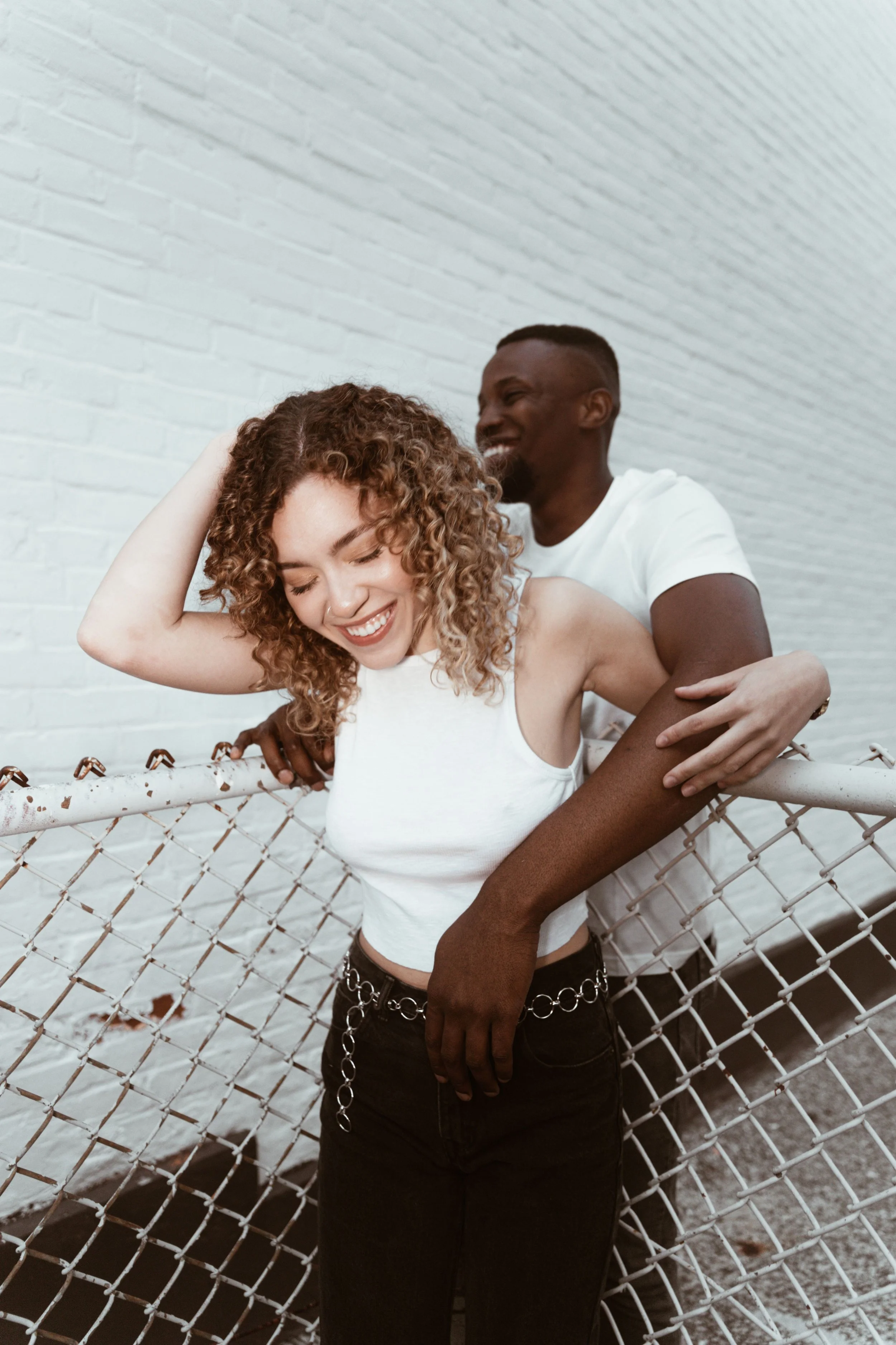 A man and woman embrace and smile against a white brick wall with a chain-link fence in the foreground.