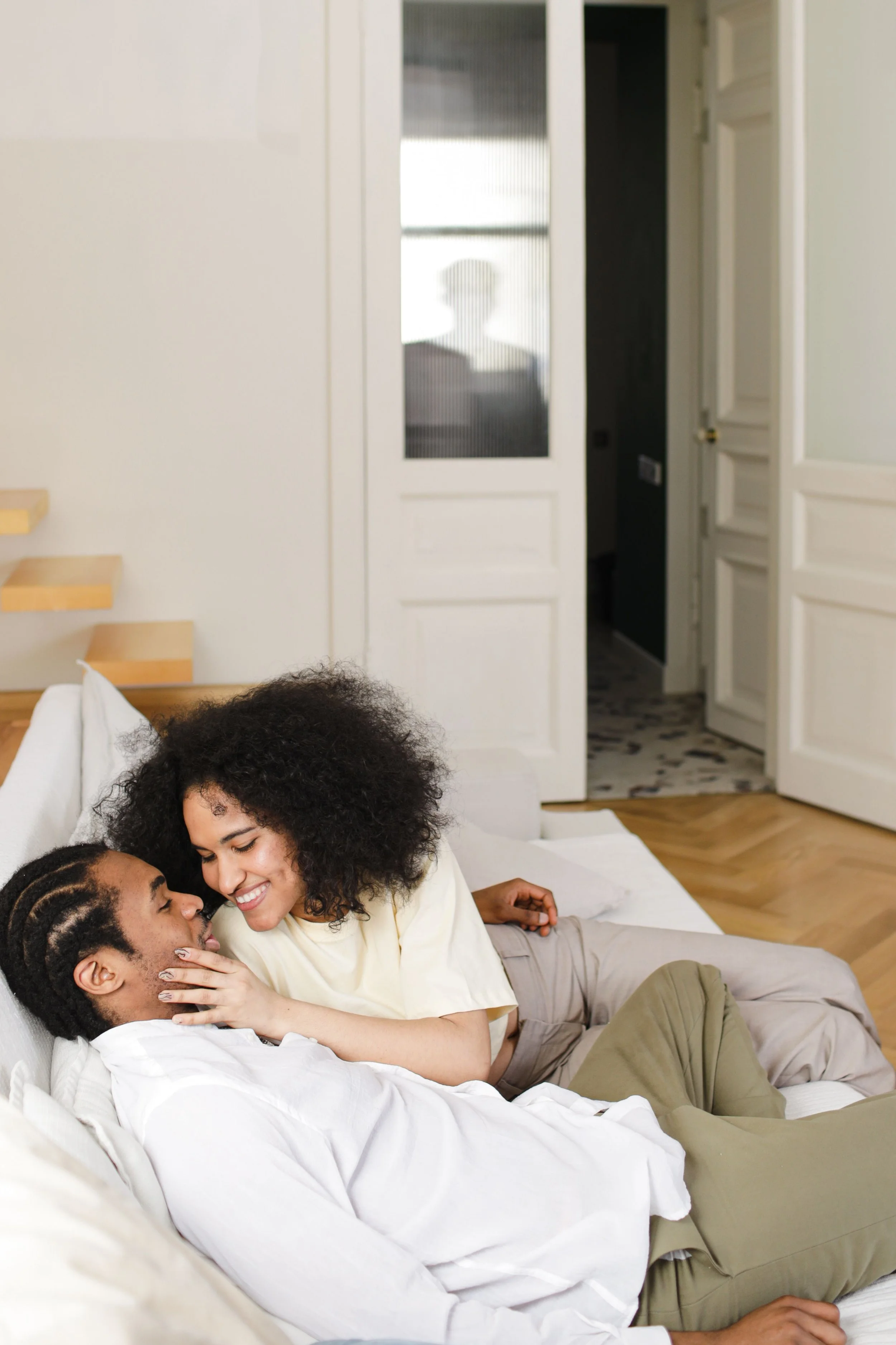 A couple lying on a bed, smiling and touching foreheads, with a woman leaning toward a man, in a cozy room.