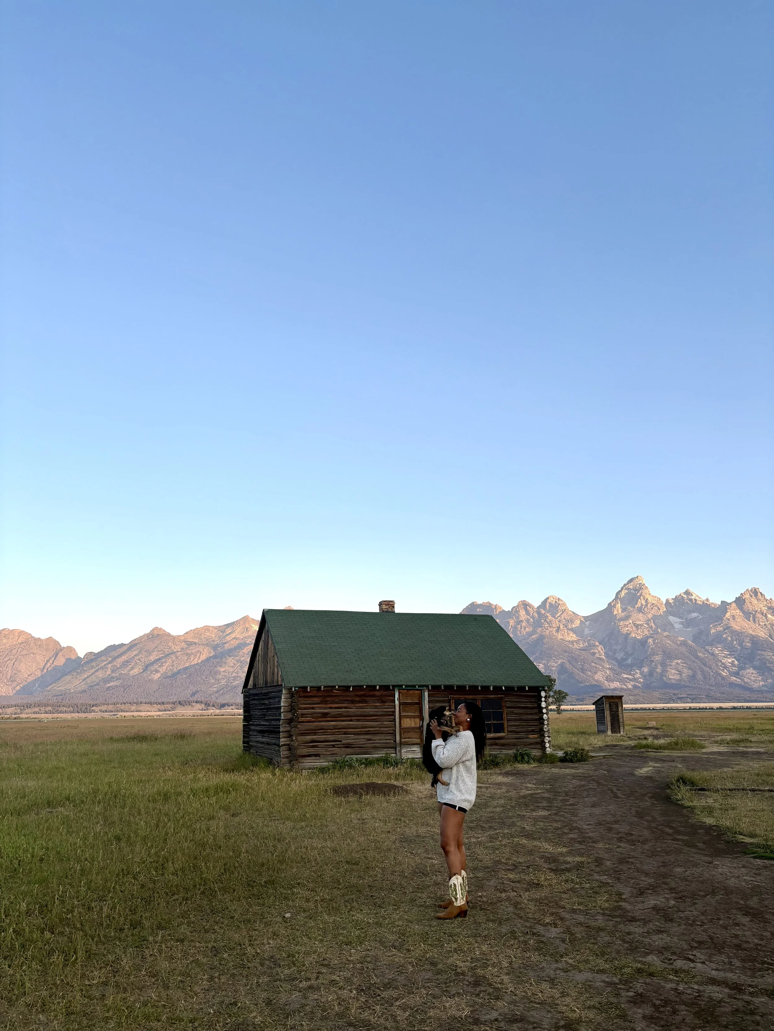 A woman in a white dress and cowboy boots holding a small dog, standing on a grassy field in front of a small wooden house. In the background, there are mountains under a clear blue sky.