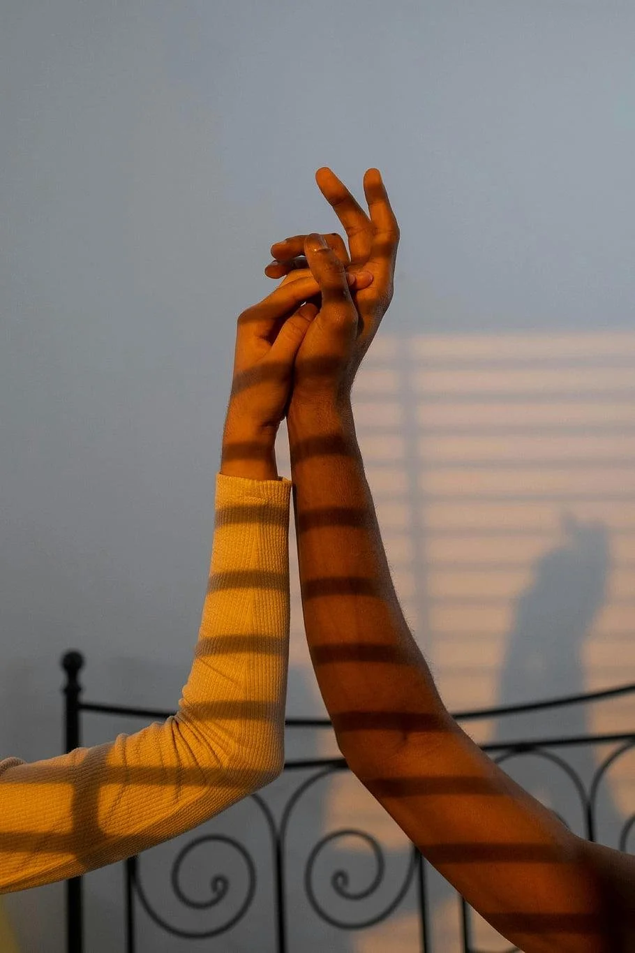 Two hands, one with a yellow sleeve, intertwine with the other, against a plain background with a black railing and soft shadows created by window blinds.