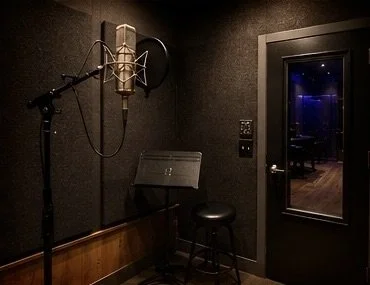 Recording studio booth with a microphone on a stand, a music stand, a stool, and a glass window showing an adjacent room.