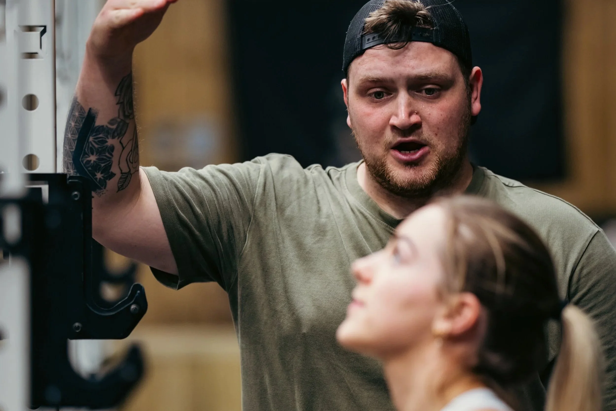 A man with a beard wearing a black baseball cap backwards, a gray shirt, and tattoos on his arm, raising his hand to adjust or hold a gym machine, while a young girl with light brown hair in a ponytail, wearing a white shirt, exercises in a gym.