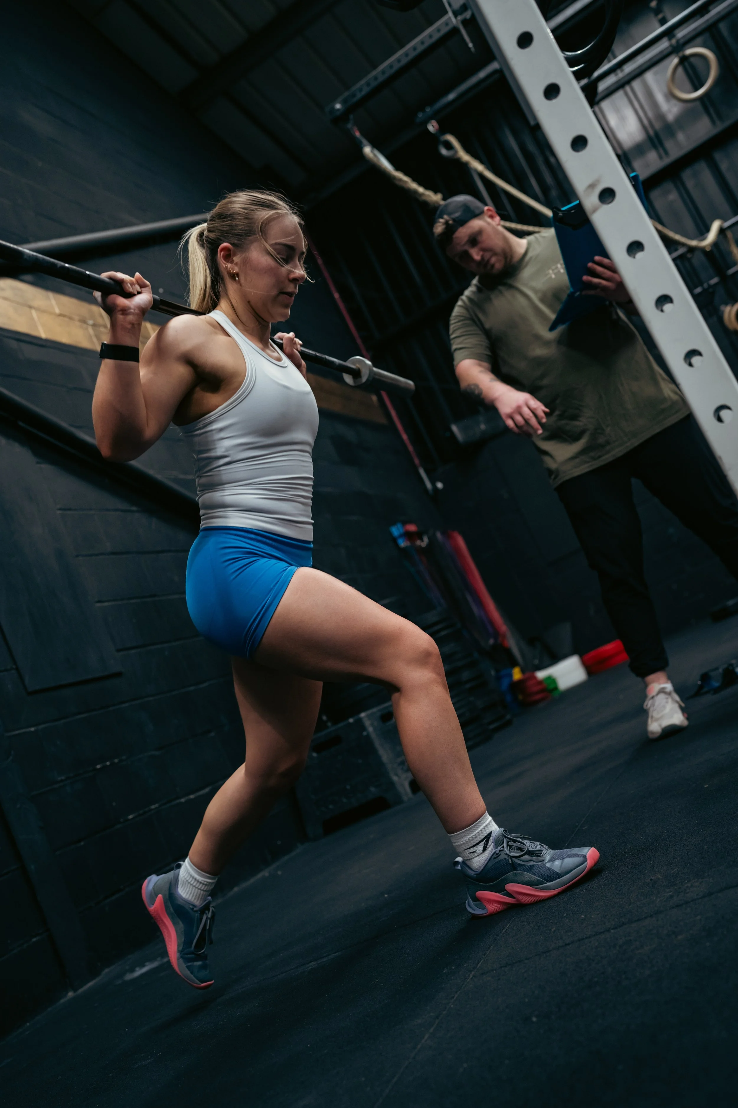 Sports therapist assessing a female athlete performing a barbell split squat during strength, movement and rehabilitation testing in a CrossFit gym.