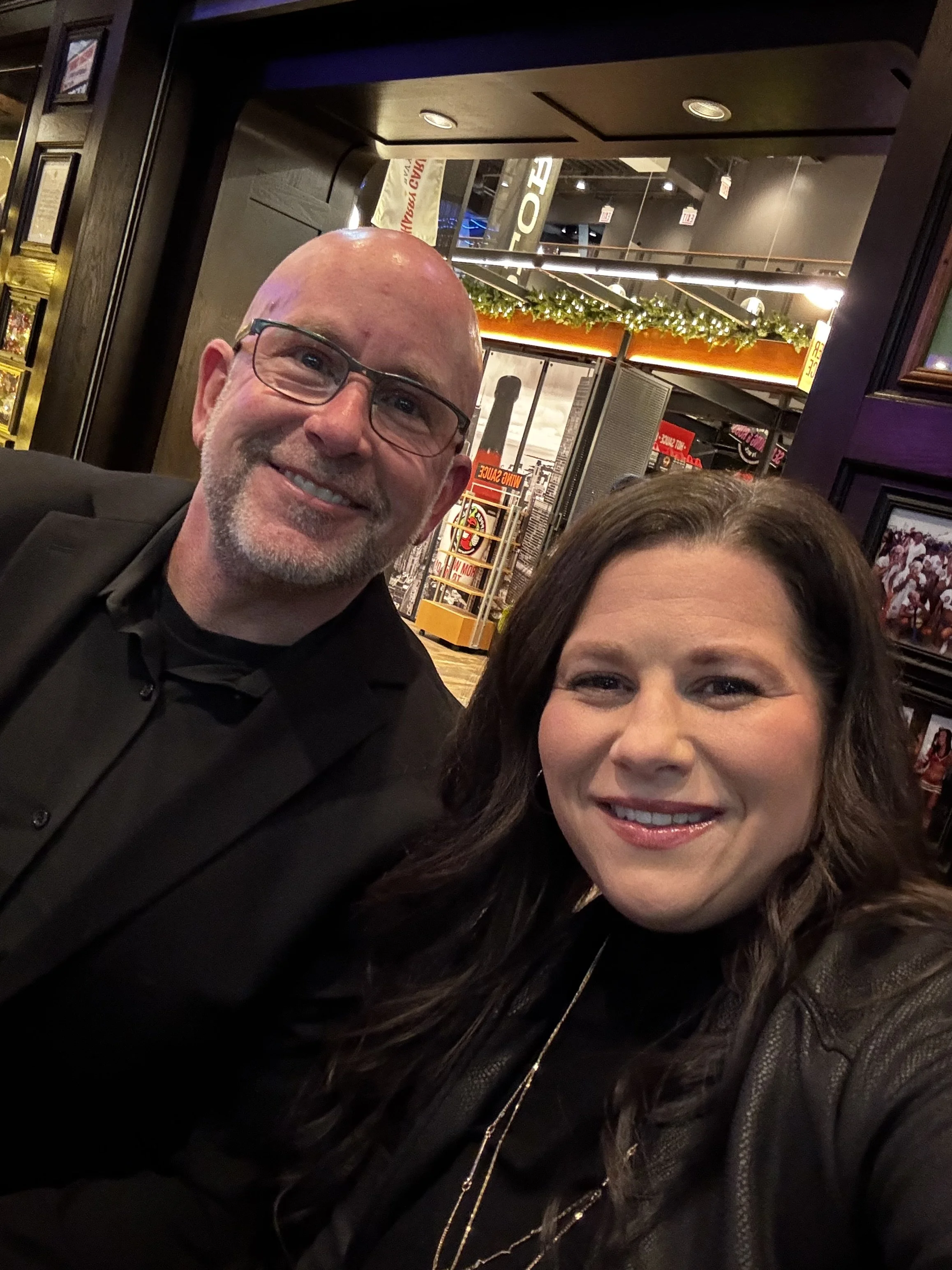 A smiling bald man wearing glasses and a woman with dark hair smiling for a photo in a restaurant or bar. The background has decorations, signs, and a warm lighting atmosphere.