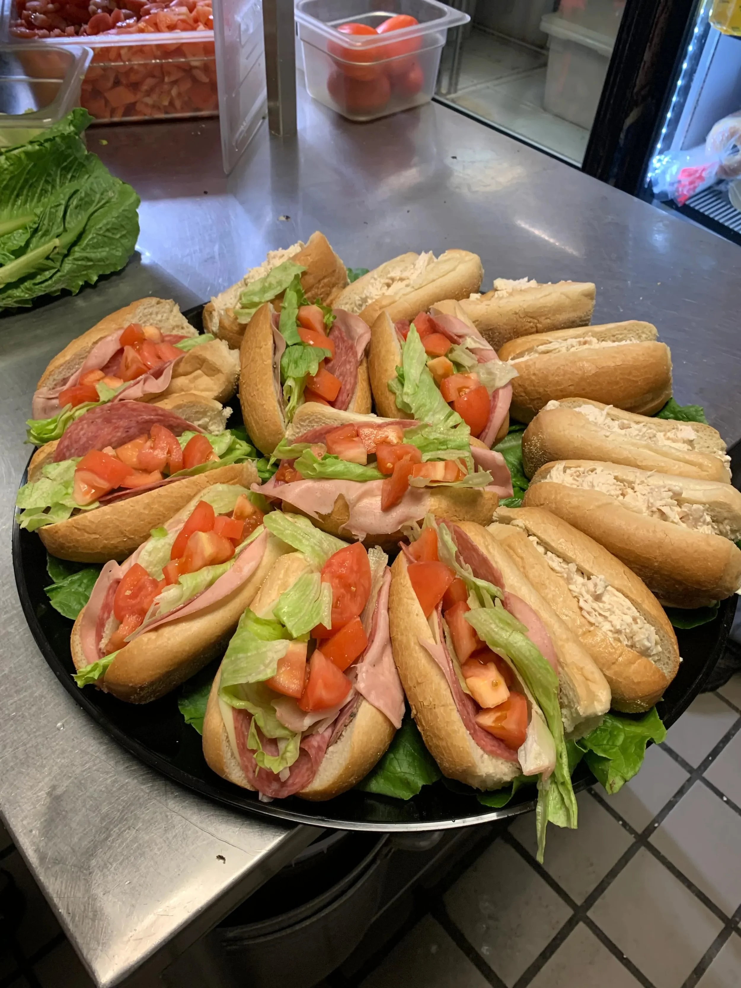 A black tray with multiple hot dogs topped with chopped tomatoes, lettuce, bacon, and other condiments. The tray is on a metallic counter with a lettuce bunch and plastic containers in the background.