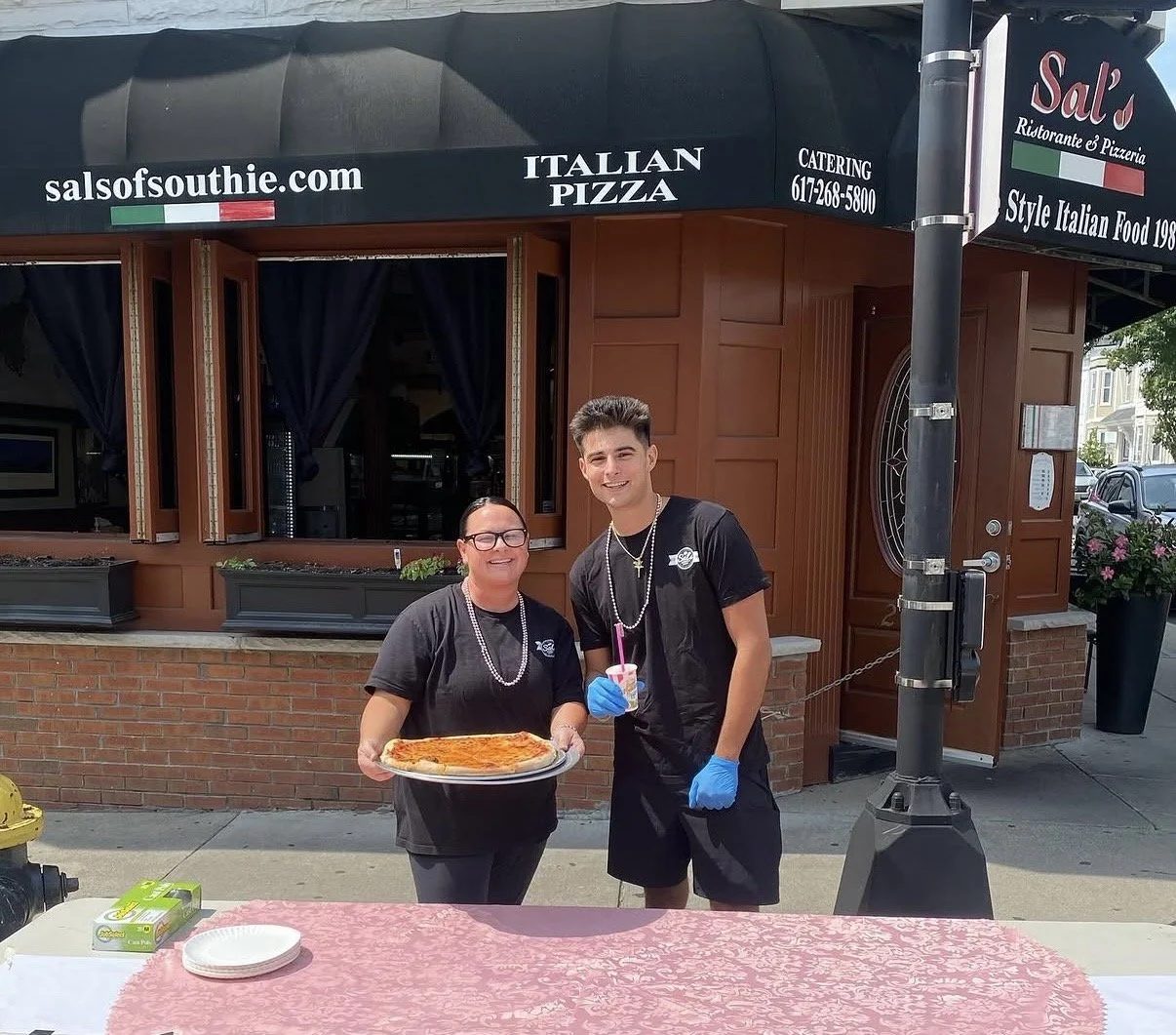 Two people standing outside a restaurant, one holding a large pizza and the other holding a drink with a straw. Both are wearing black shirts and blue gloves, smiling at the camera. The restaurant sign reads 'Sal's Italian Pizza' and 'salsofsouthie.c