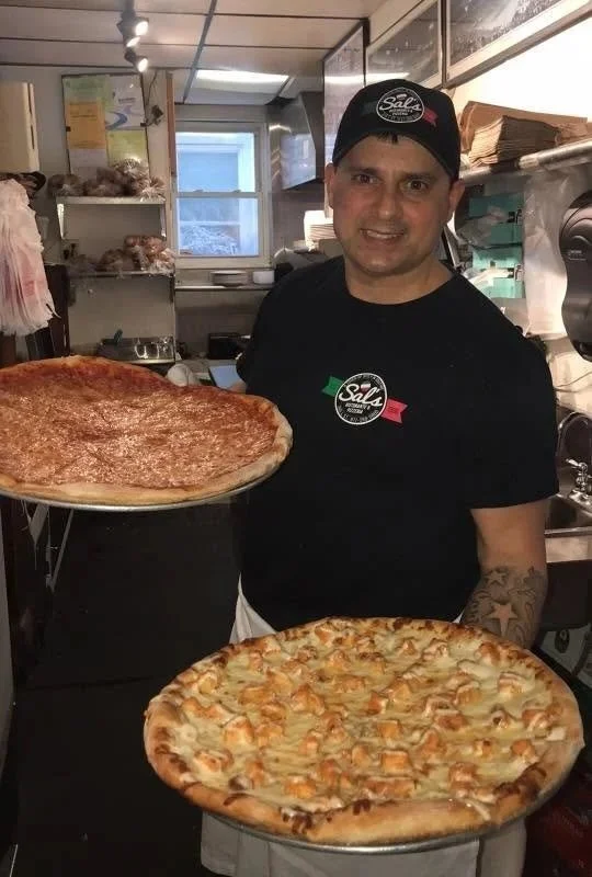 A man in a black uniform holding a large pepperoni pizza, standing in a kitchen with another large cheese pizza on a table in front of him.