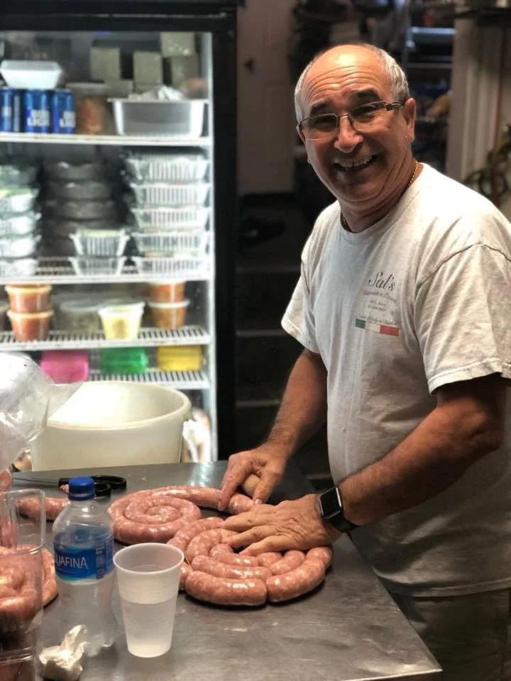 A man smiling while preparing raw sausages on a kitchen counter, with a refrigerator in the background.