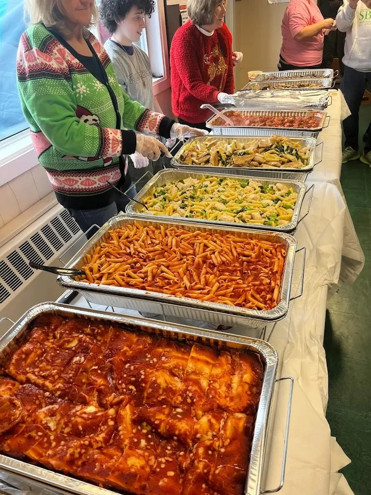 A line of people serving food at a buffet table filled with trays of pasta and Italian dishes, including penne pasta with marinara sauce, chicken and vegetable pasta, and other cooked dishes.
