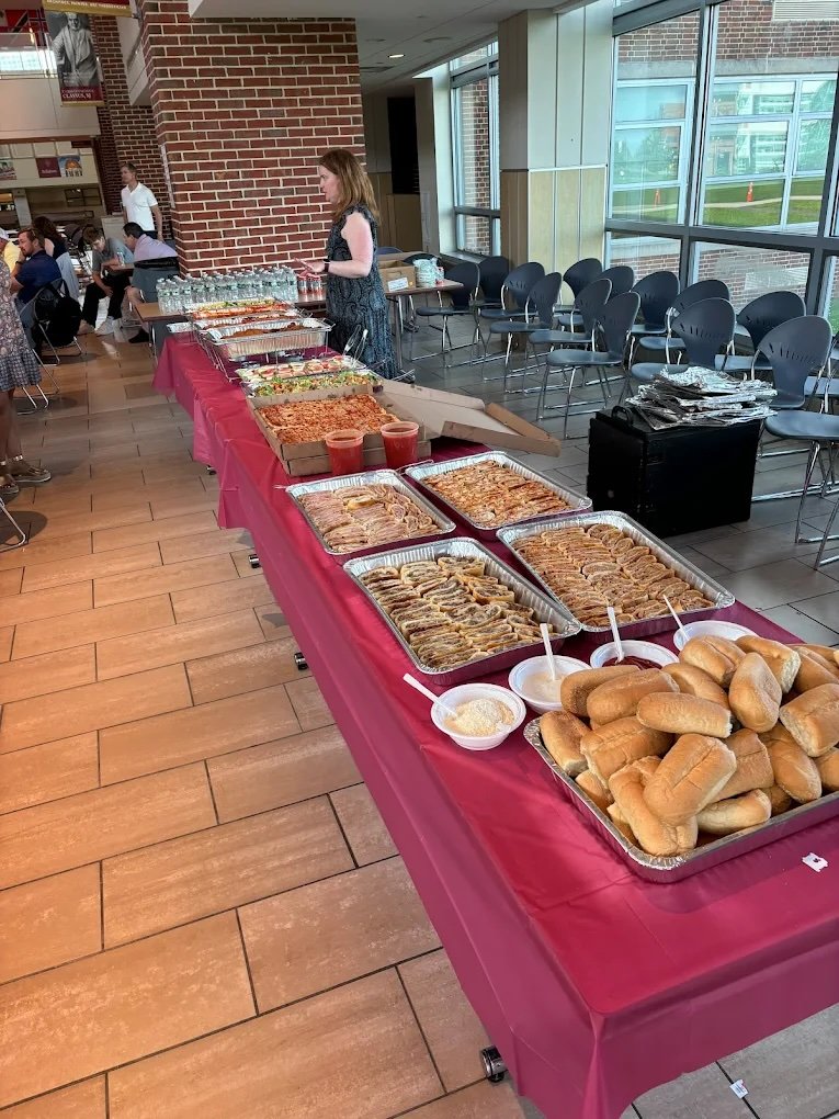 Buffet table with hot dog buns, pizza, fried food, and condiments set up for a gathering or event in a spacious room with large windows and seating area.