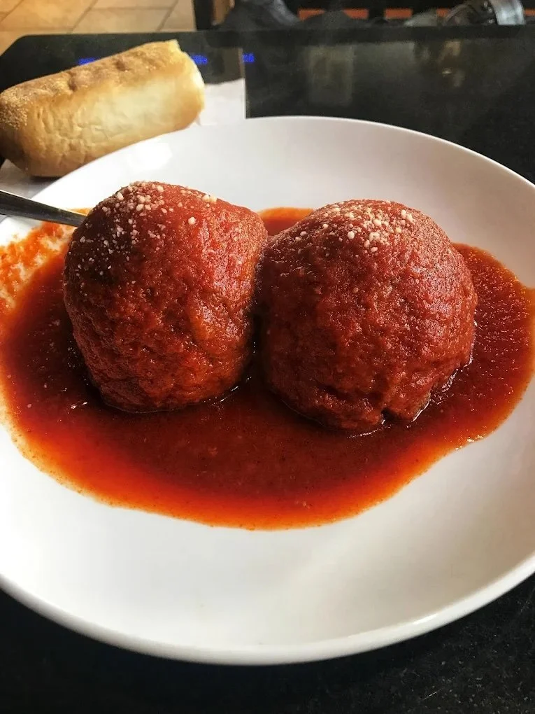 Two Italian meatballs in marinara sauce on a white plate with a piece of garlic bread in the background.