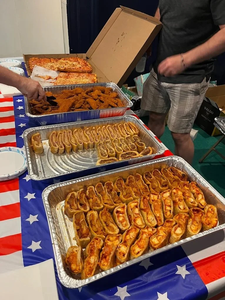 Buffet table with cheese pizza, fried chicken wings, and assorted mini sandwiches, decorated with American flag-themed tablecloth, at casual gathering.