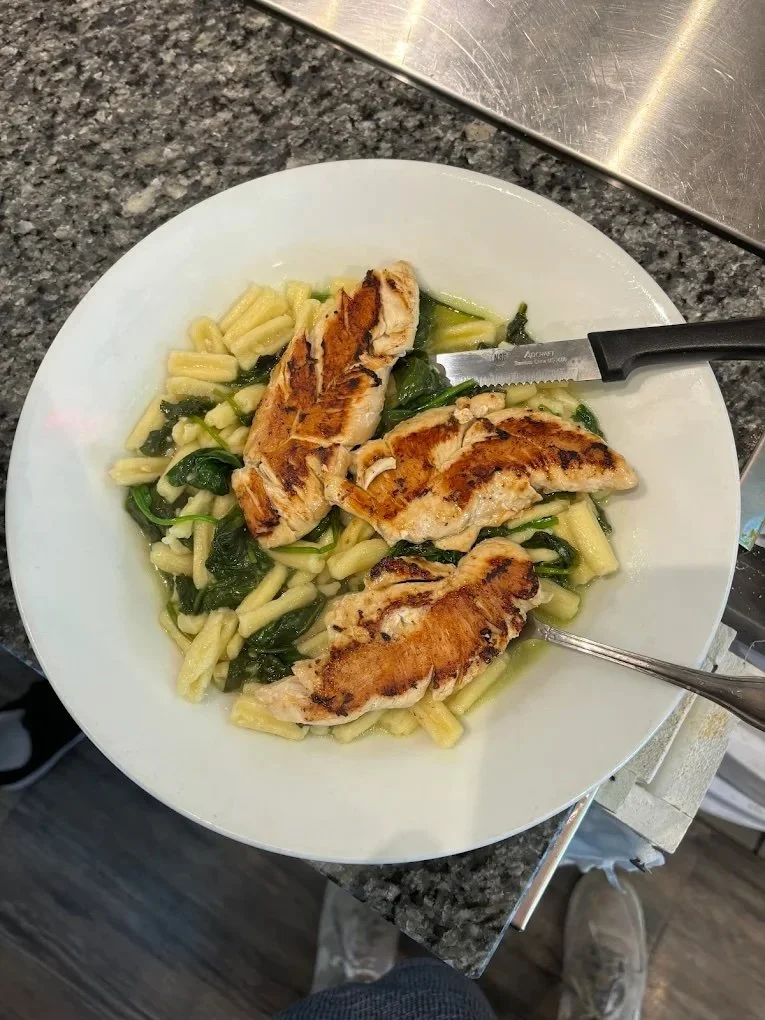 Grilled chicken breast served on pasta with spinach on a white plate.