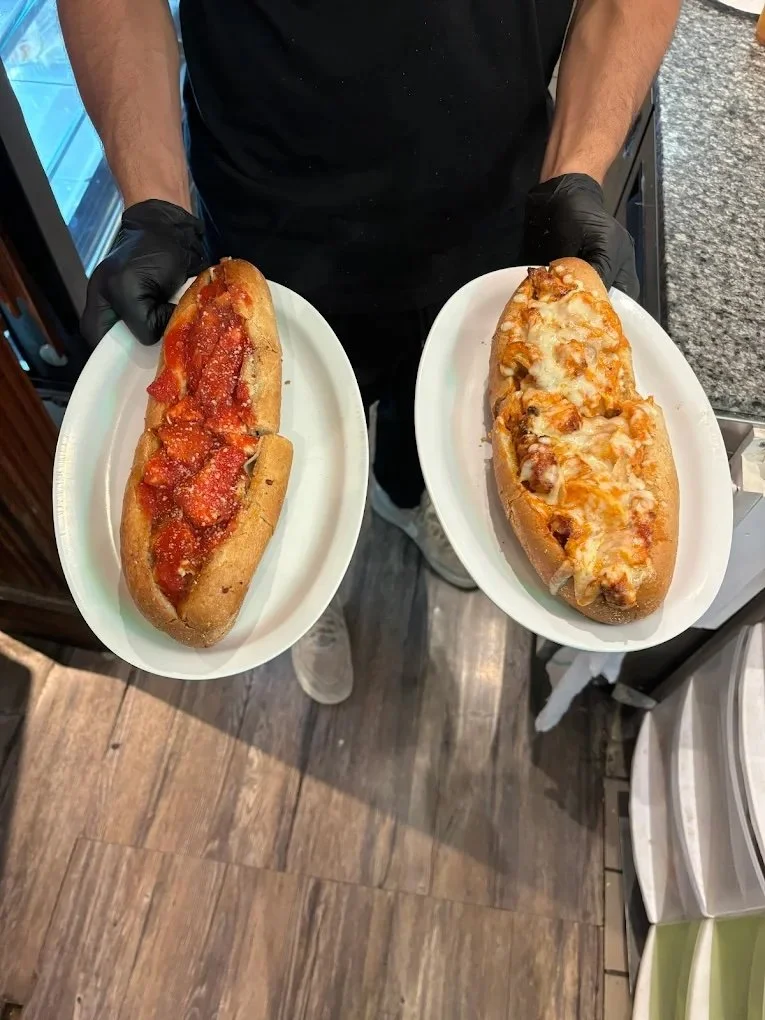 A person wearing black gloves holding two plates each with a large Italian sub sandwich, one with pepperoni and tomato sauce, the other with cheese and melted toppings.