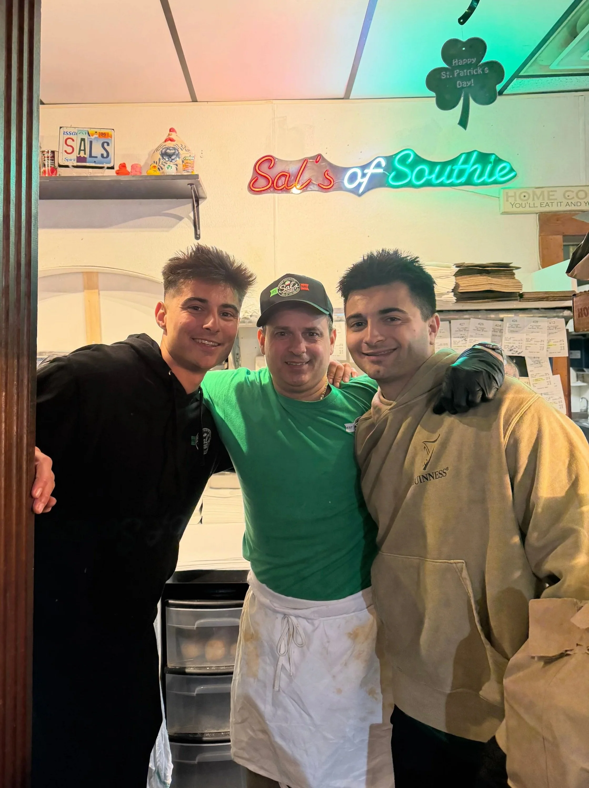 Three men standing inside a restaurant, smiling at the camera, wearing casual jackets and an apron. The background features a neon sign that reads 'Sal's of Southie', a shamrock decoration with a sign saying 'Happy St. Patrick's Day!', and shelves with papers and packages.