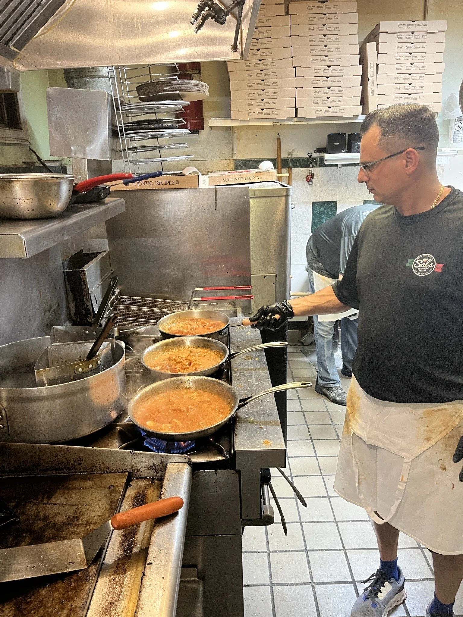 A man cooking in a restaurant kitchen, wearing glasses, a black T-shirt, a white apron, and black gloves. He is stirring a pot of soup or stew on a stovetop. Behind him are stacks of pizza boxes and kitchen utensils.