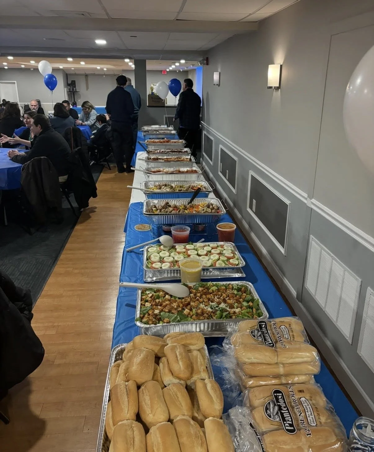 A buffet table with various dishes including bread rolls, sandwiches, salads, and hot foods at an indoor event with guests seated at tables in the background.