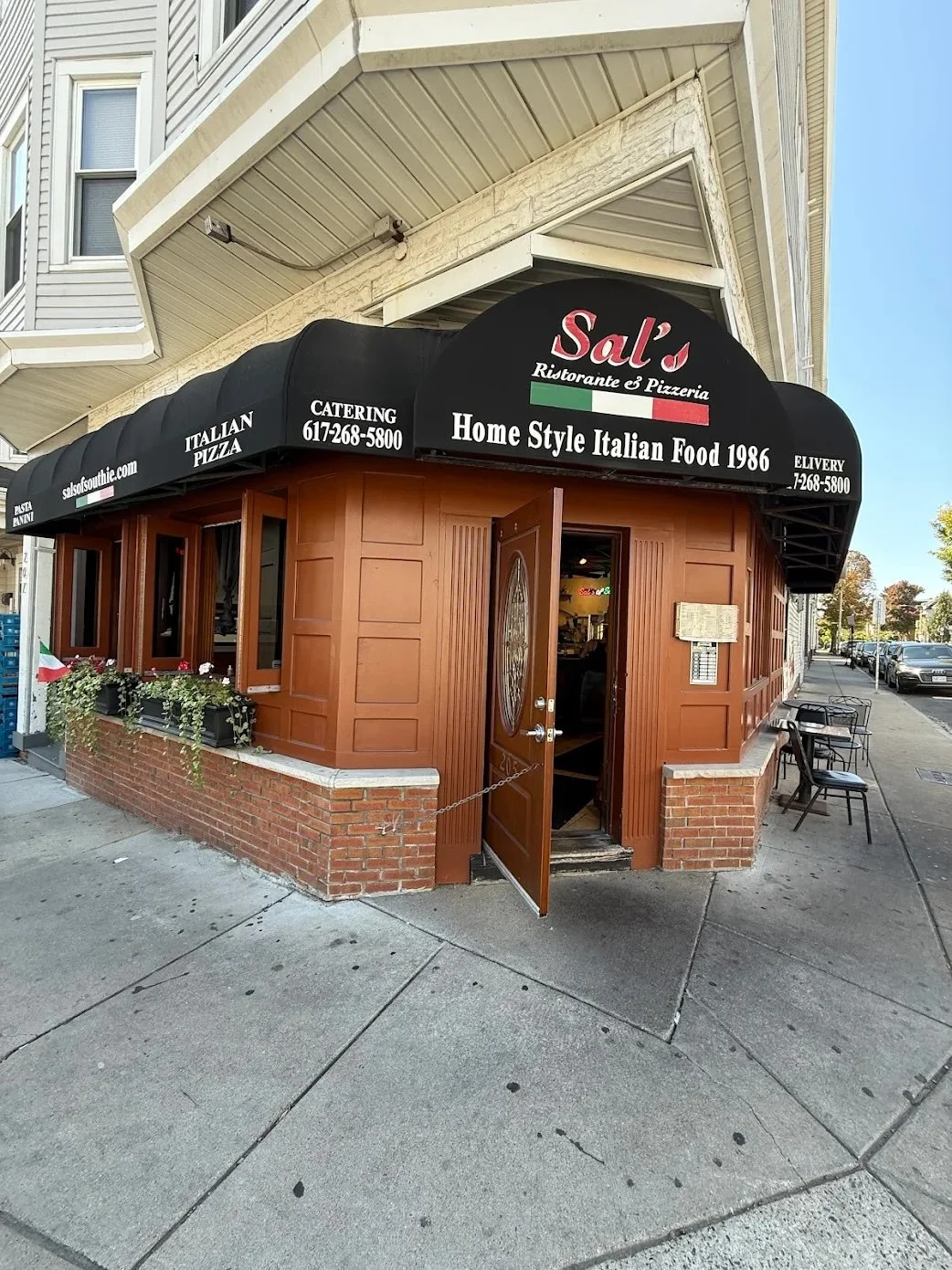 Corner of a small Italian restaurant with a black awning that reads "Sal's Ristorante & Pizzeria, Home Style Italian Food 1986". The building has wooden paneling, a brick foundation, and an open door. Outdoor seating with tables and chairs is visible on the sidewalk.