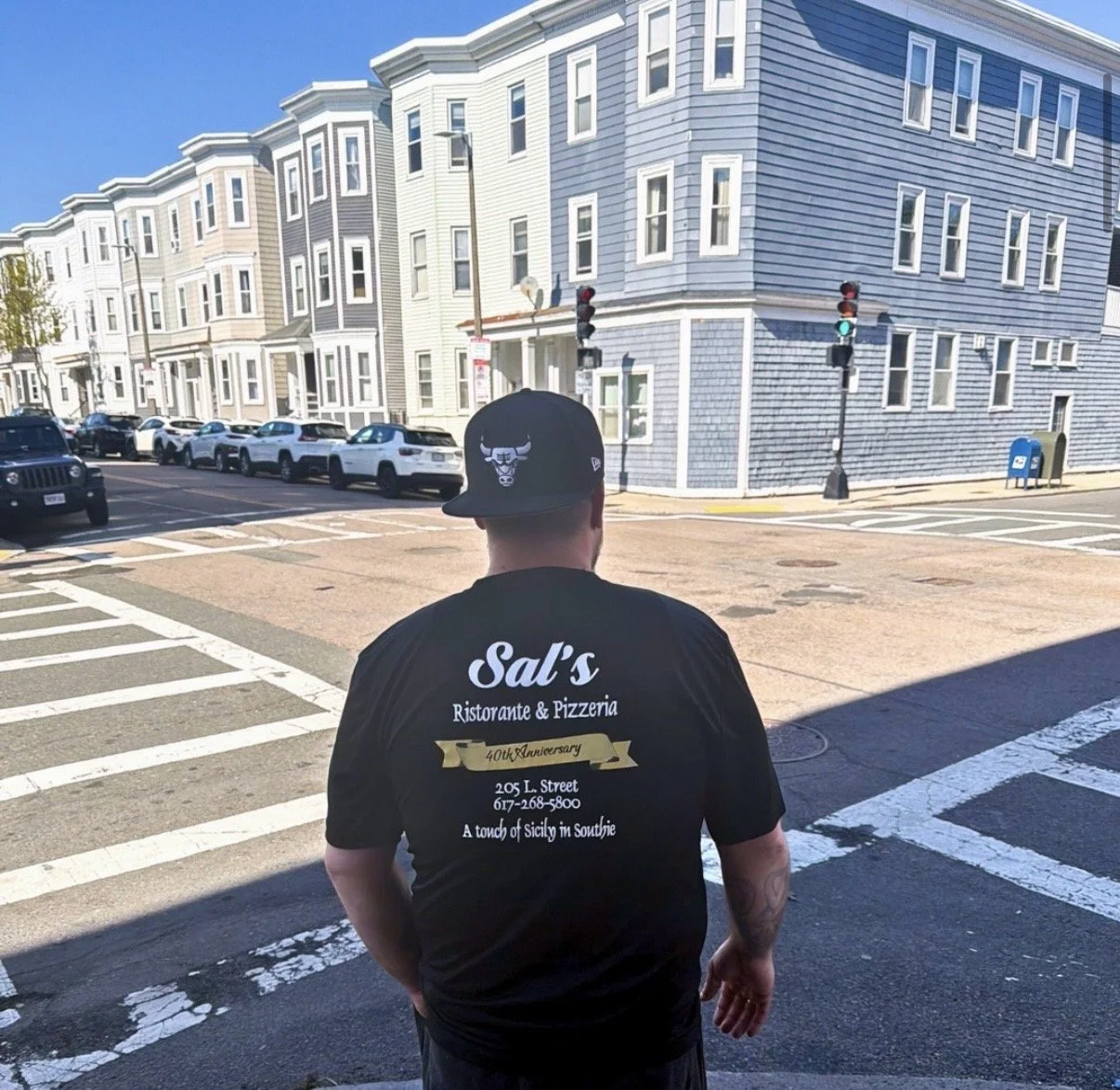 A man wearing a black Chicago Bulls cap and a black T-shirt with a 40th anniversary Sals of Southie tee stands at a crosswalk in South Boston with parked cars and a row of pastel-colored residential buildings on a sunny day. 