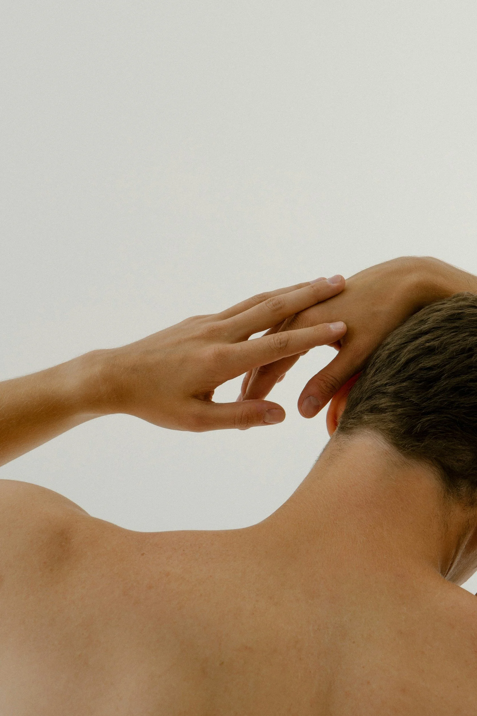 Close-up of a person with short hair, bare shoulder, and skin, covering one eye with their hand, against a plain white background.