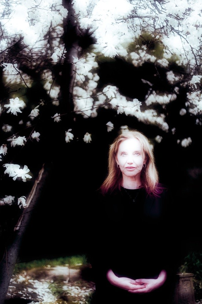 A woman with light brown hair standing in front of a flowering tree with white blossoms, partially illuminated, with her hands clasped in front of her.