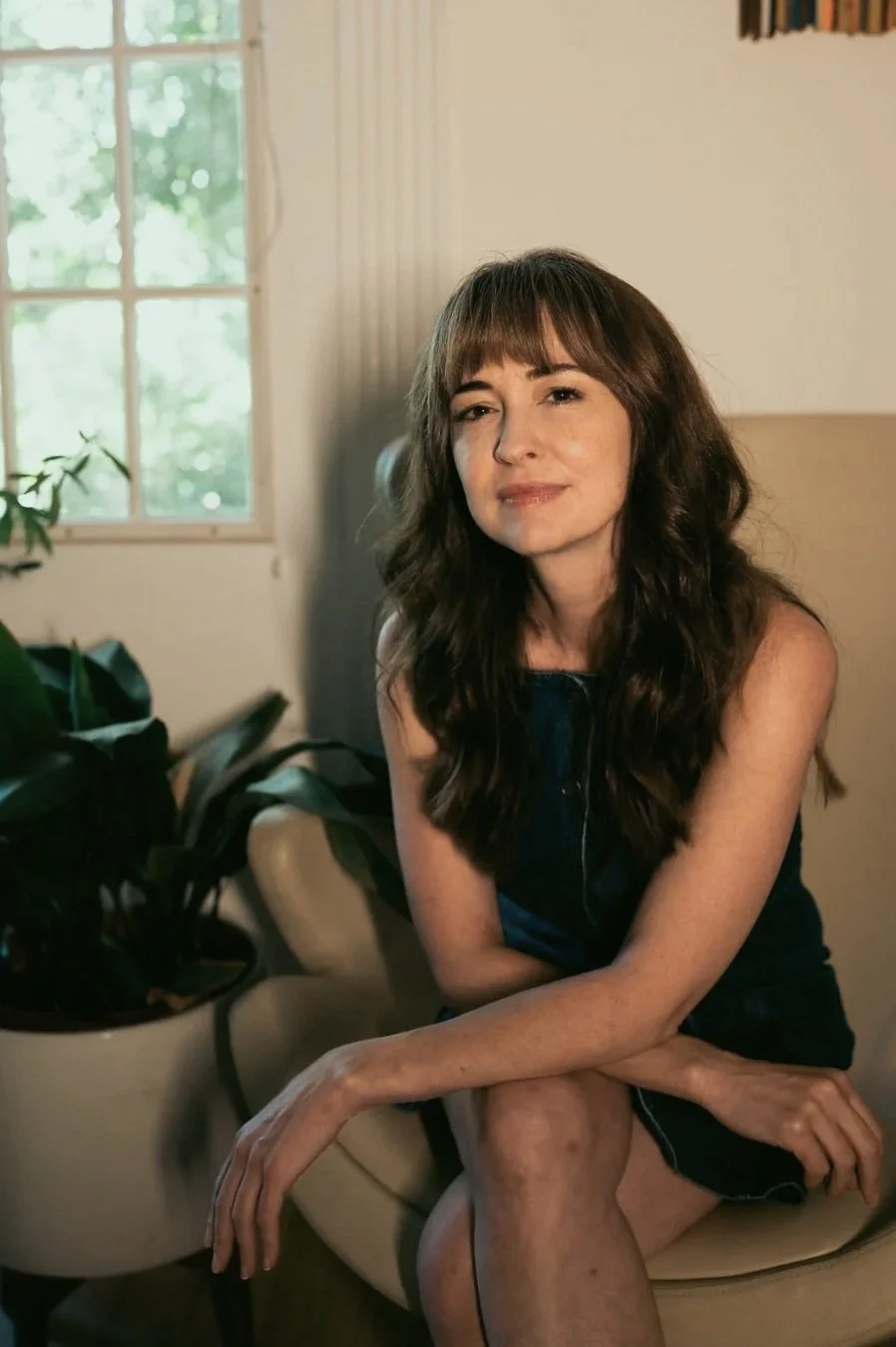 A woman with long wavy brown hair sitting on a beige couch in a room with a large window and a plant nearby.