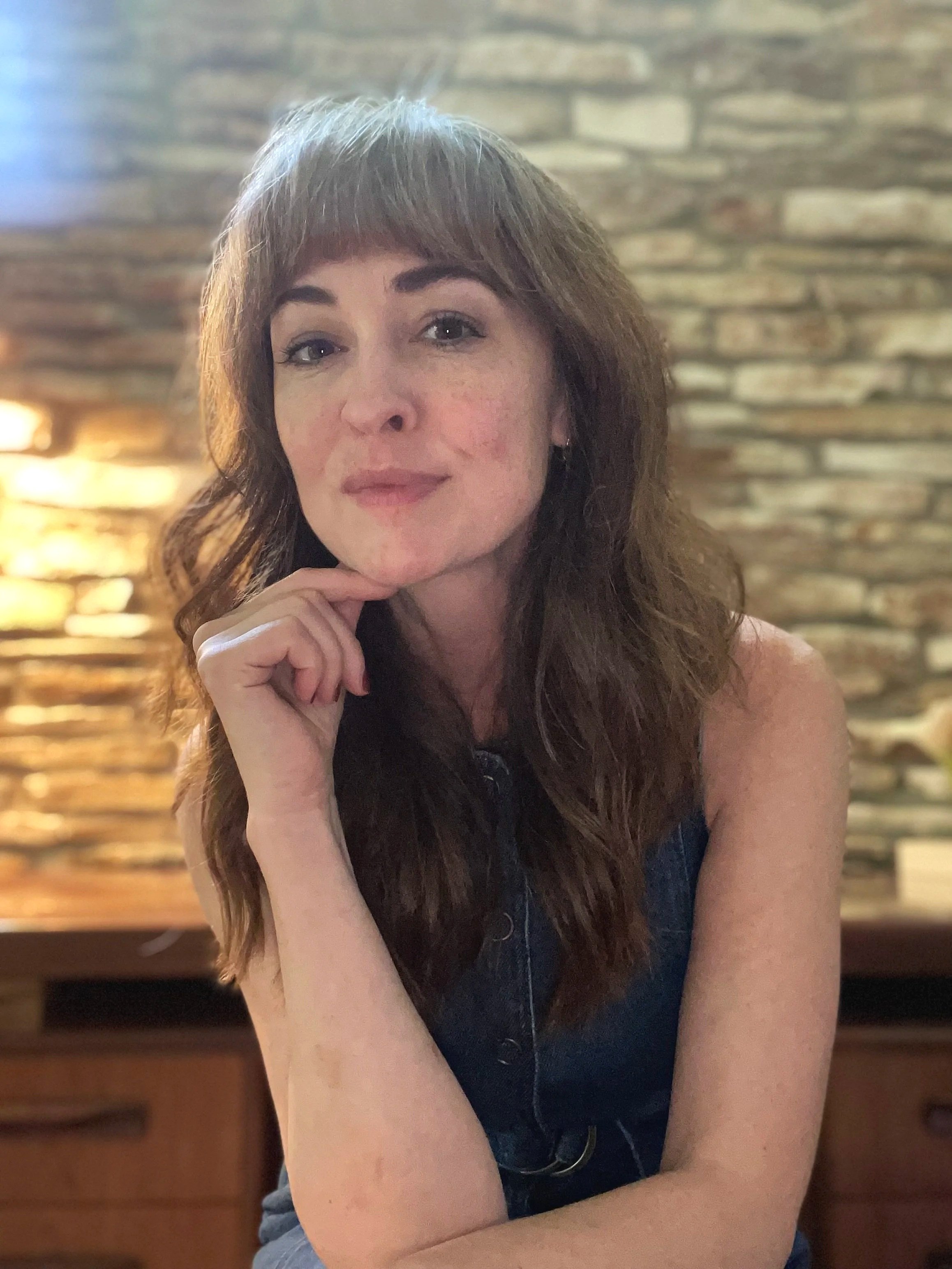A woman with reddish-brown hair and bangs is posing indoors with a brick wall background. She has her chin resting on her hand, and she is wearing a sleeveless top.