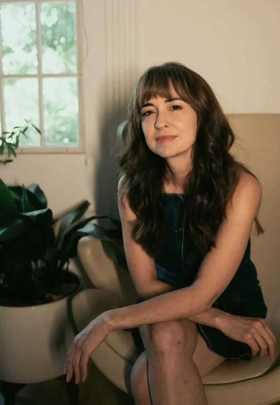 A woman with long wavy brown hair sitting on a beige chair indoors near a window, with a plant nearby, wearing a dark sleeveless dress, looking at the camera with a slight smile.