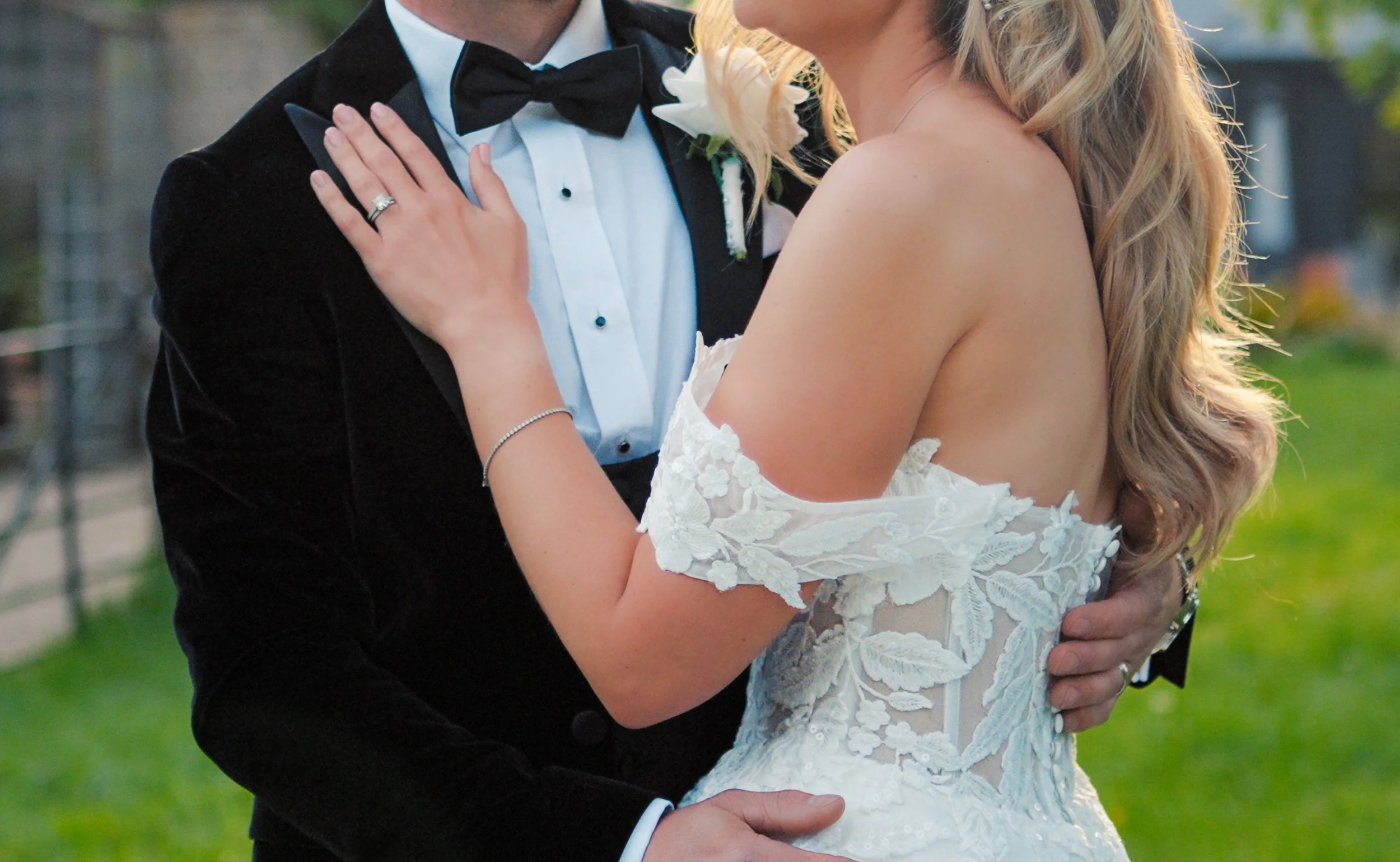 Close-up of a bride and groom hugging outdoors, the bride wearing a white lace wedding dress with floral details on the sleeves, and the groom in a black tuxedo with a bow tie and a white boutonniere.